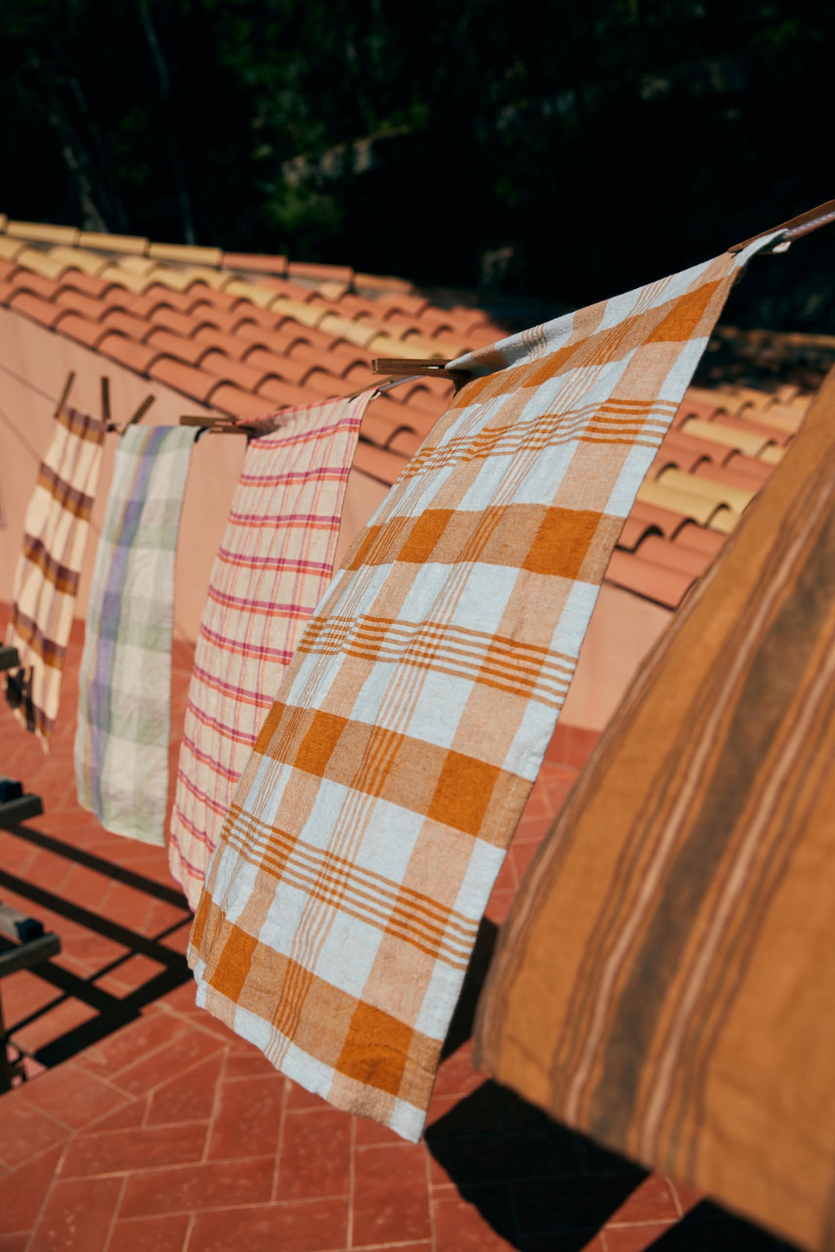 The HKLIVING Vida Check Tea Towel in Strawberry/Cream hangs on a clothesline above a tiled terrace, with a terracotta roof in the background.