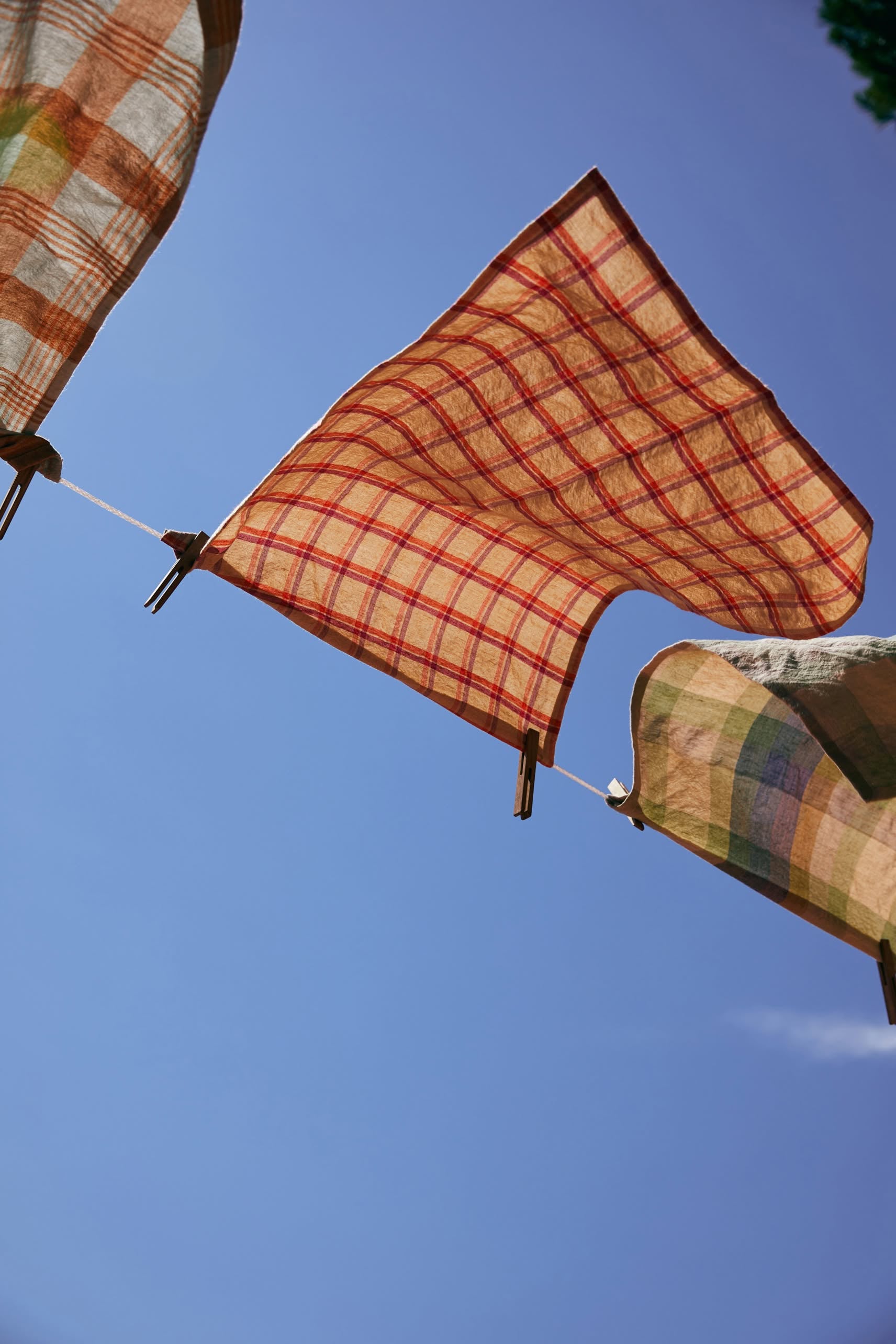The HKLIVING Vida Check Tea Towel in Strawberry/Cream, made of linen and cotton, hangs fluttering on a clothesline against a clear blue sky.