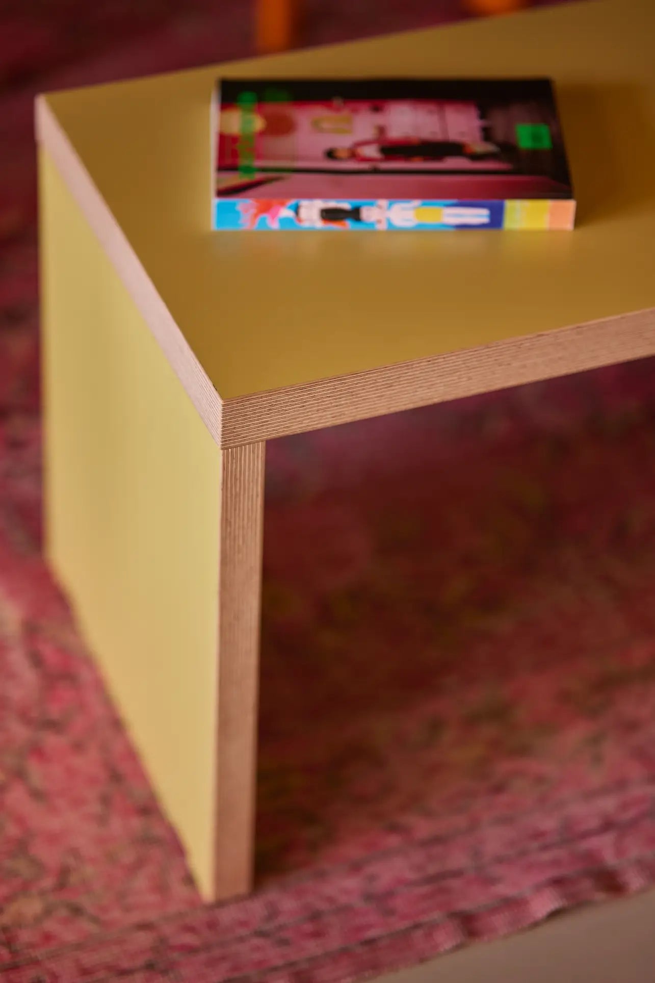 A close-up of a yellow wooden dining table with a colorful book on top, set on a pink patterned rug—ideally complemented by the HKliving Bench, 220cm from HKliving for stylish, space-saving seating.