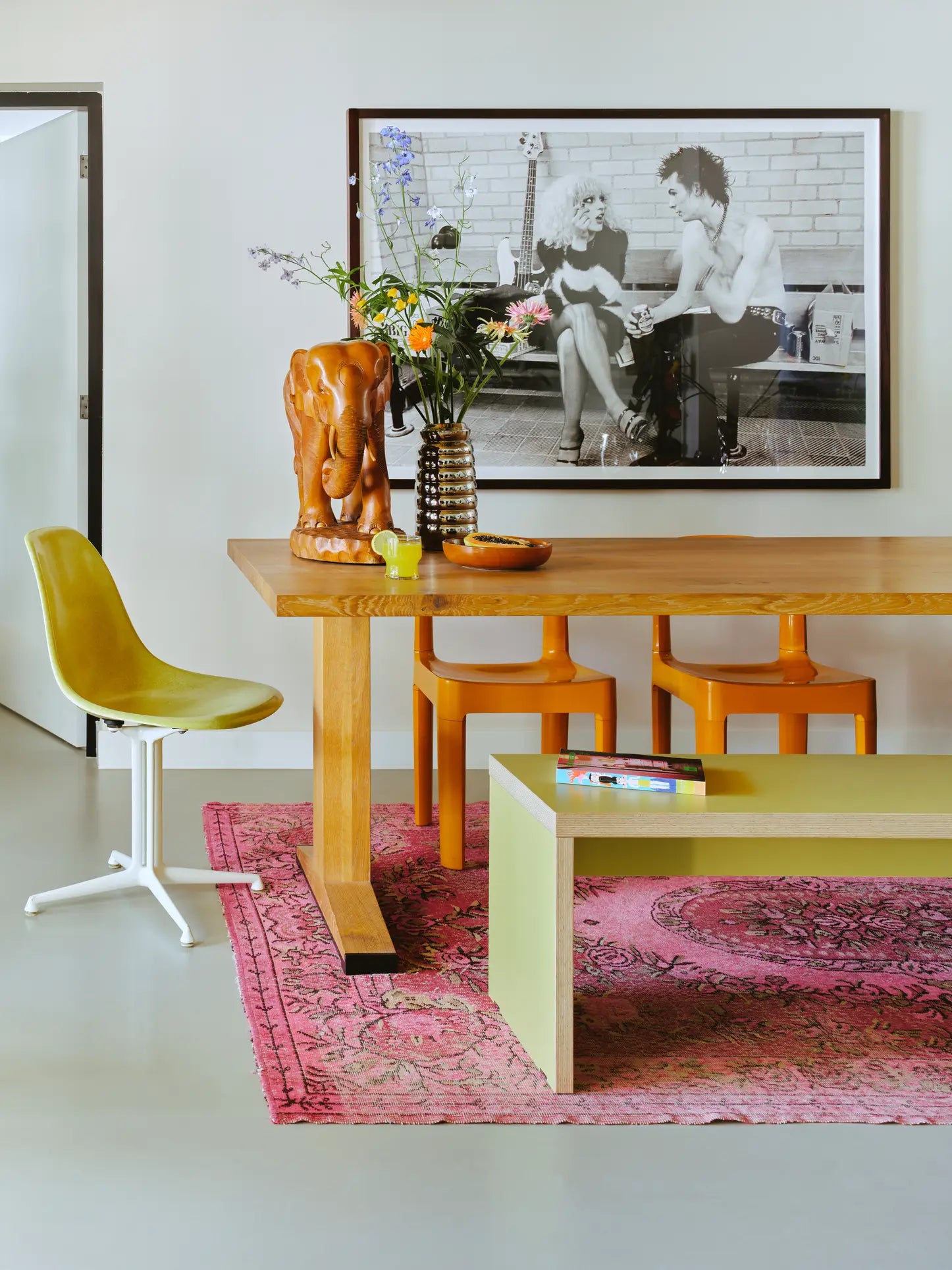 A modern dining space with a space-saving wooden table, yellow chairs, a white-legged green chair, the 220cm HKliving Bench in green by HKliving, and a pink rug. Flowers and an orange elephant sculpture sit on the table; a black-and-white photo hangs nearby.