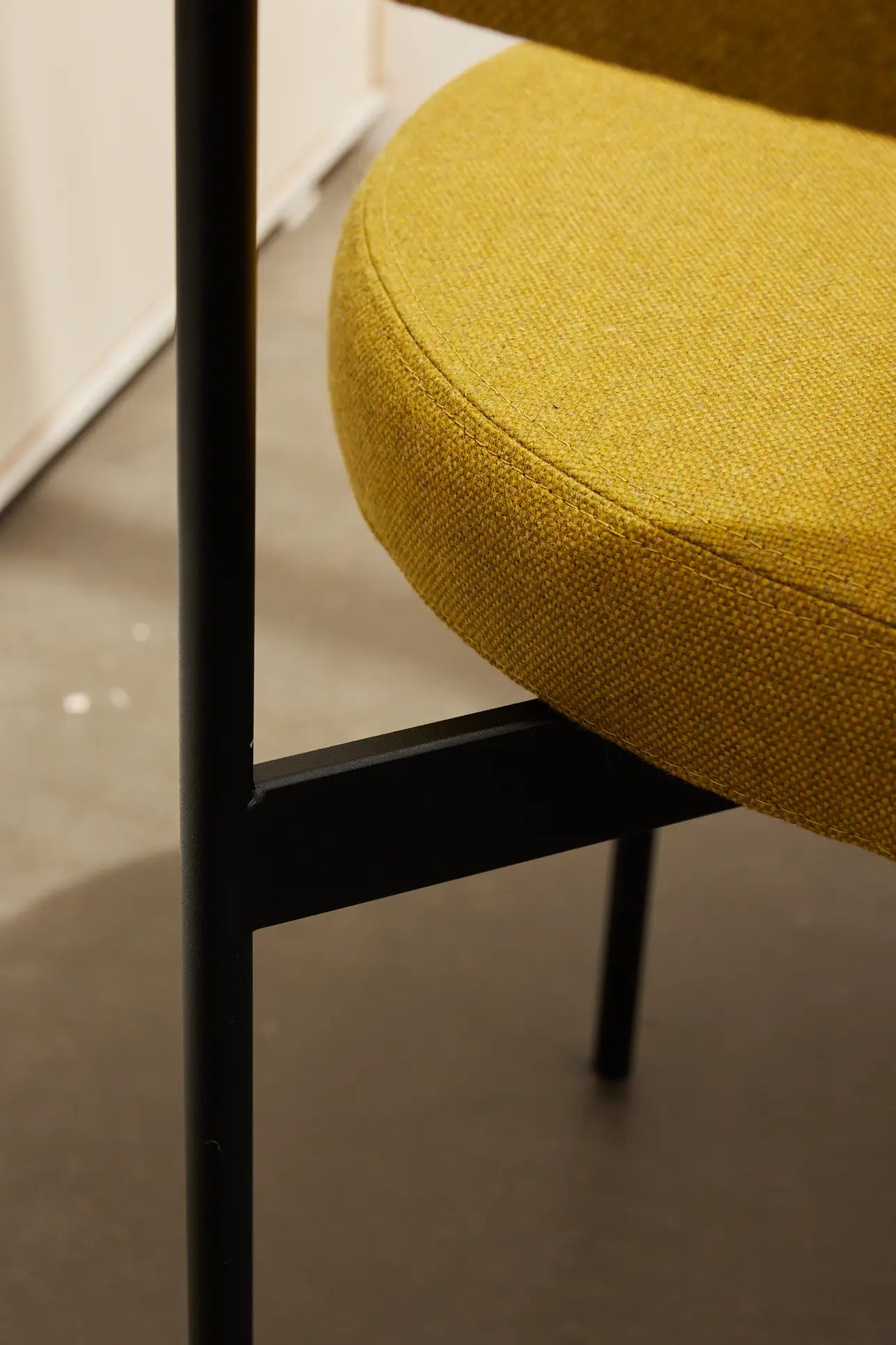 Close-up of the HKliving Dining Armchair by HKliving, inspired by Dutch design, with a round Main Line Flax mustard yellow fabric seat and black metal legs, set on a smooth gray floor.