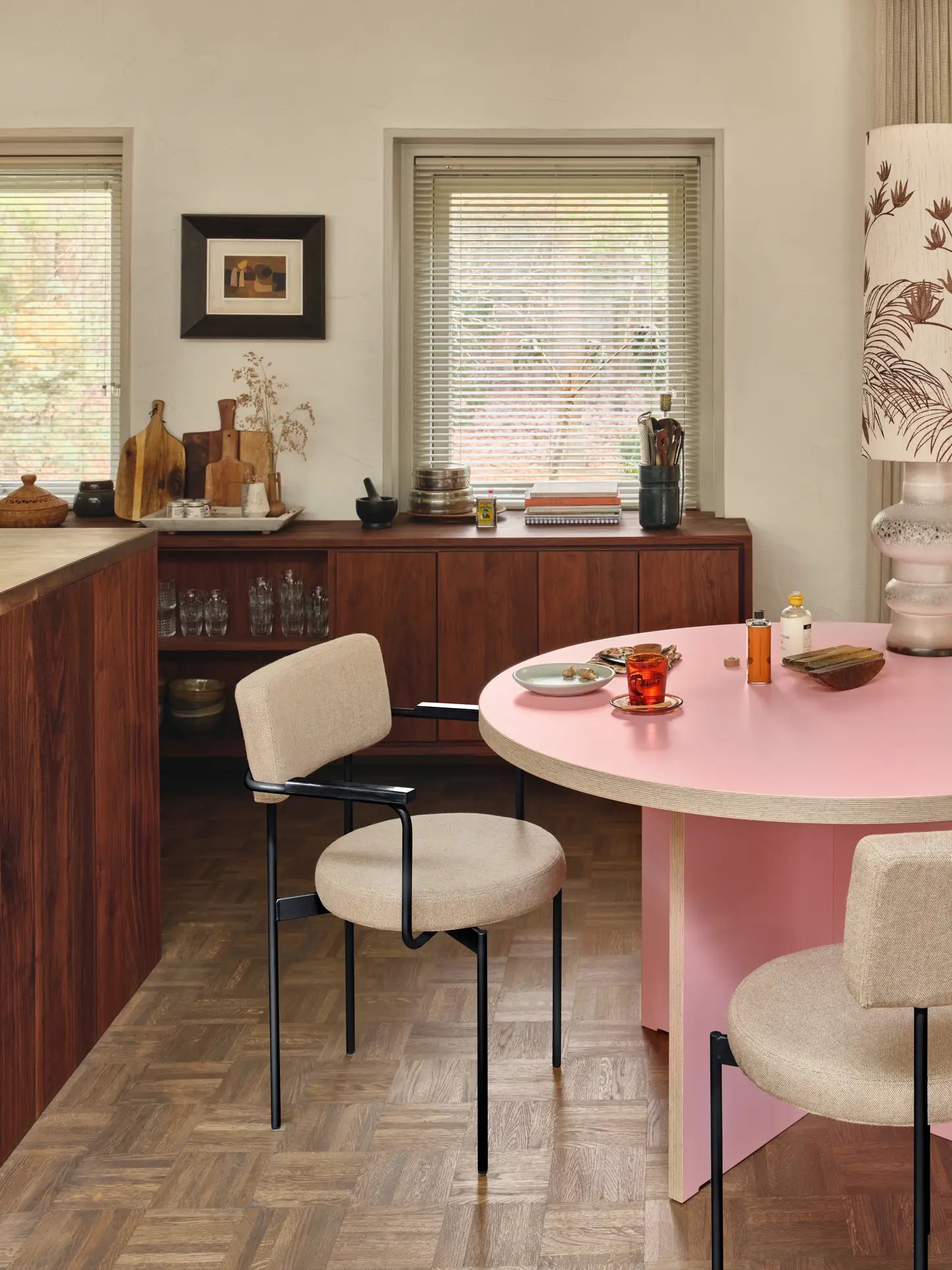 A modern kitchen features wood cabinets, a round pink table with mugs and a vase, HKliving Dining Armchairs in Main Line Flax with black legs, wooden flooring, and two windows with blinds allowing in soft natural light.