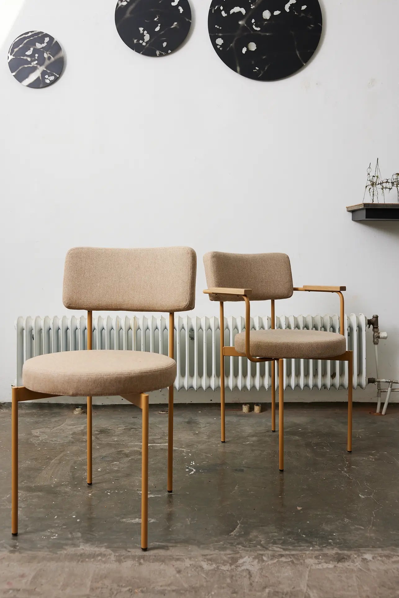 Two HKliving Dining Armchairs, Main Line Flax Dusty, with gold metal legs sit on a concrete floor by a white radiator. Behind them is a white wall displaying three round black and white abstract art pieces.