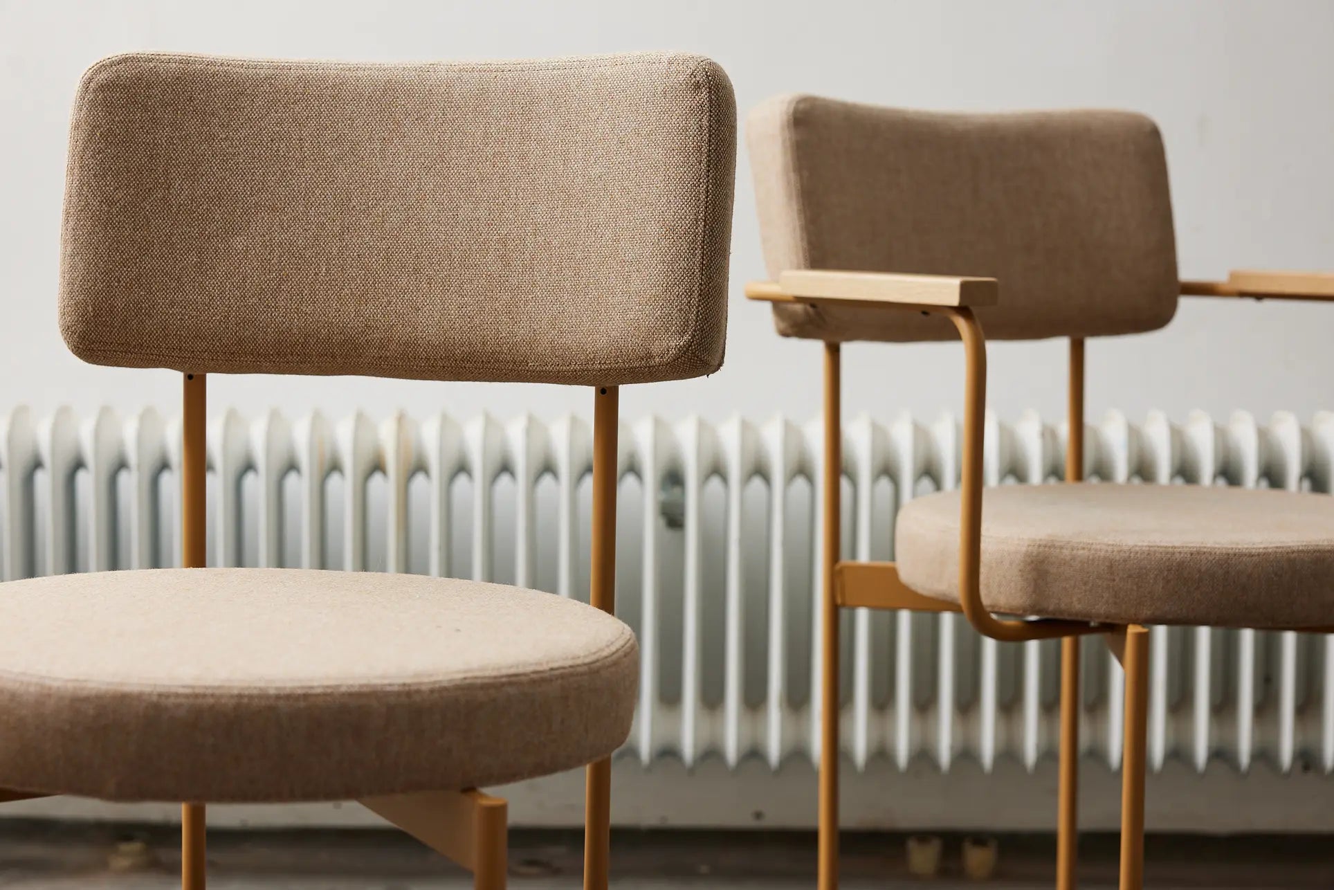 Two HKliving Dining Armchairs in Main Line Flax Dusty fabric, featuring metal frames and Dutch-inspired design, sit in front of a white radiator. The stackable beige chairs offer clean lines; one features convenient armrests.
