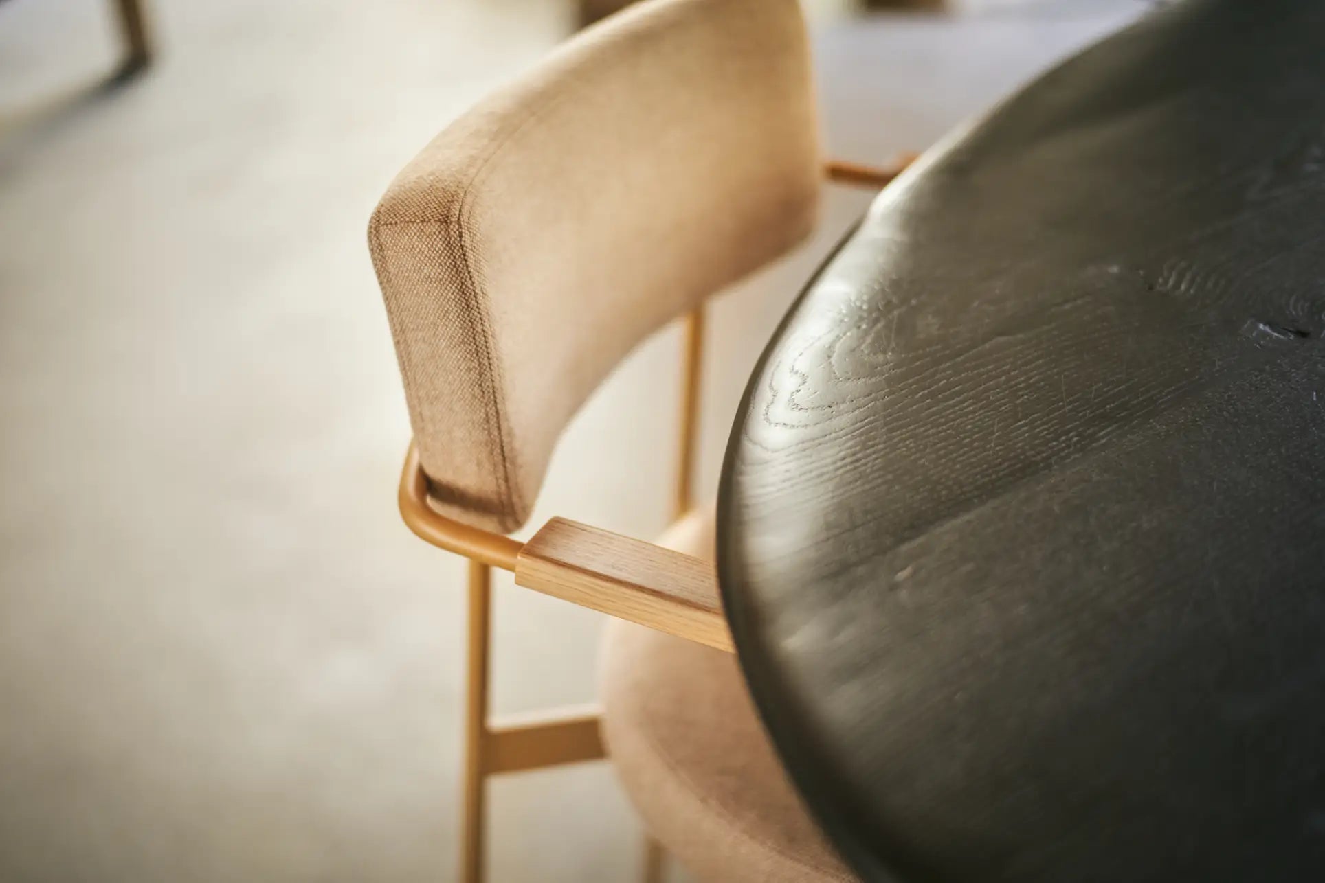 A close-up of the HKliving Dining Armchair in Main Line Flax Dusty, a modern beige upholstered seat with wooden legs, sits next to a dark round wooden table in a softly lit room.