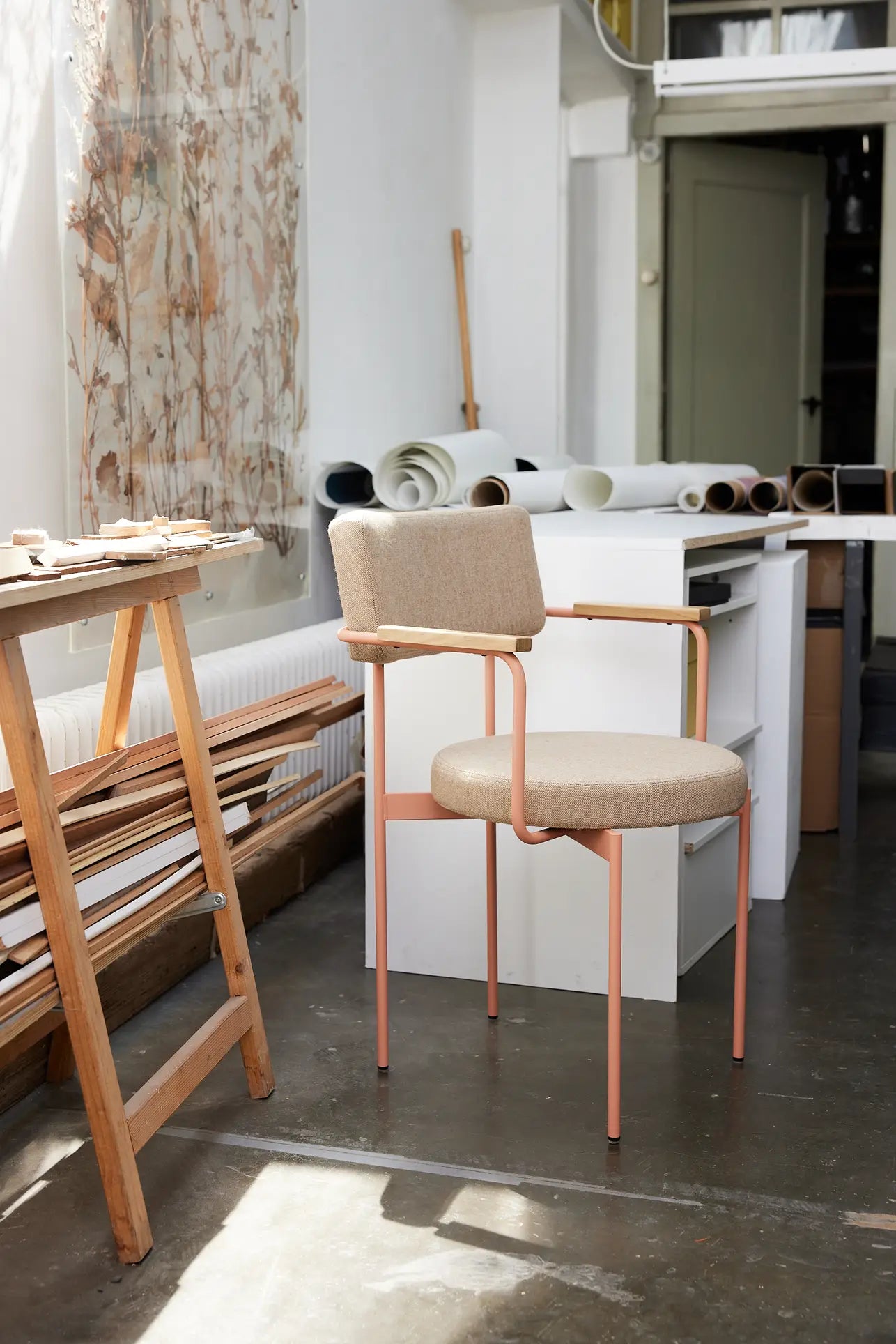 The HKliving Dining Armchair, Main Line Flax, Nude by HKliving features a beige cushion and pink metal frame, inspired by Dutch design, shown in a bright studio with wooden planks, a white desk with plans, and sheer floral curtains.