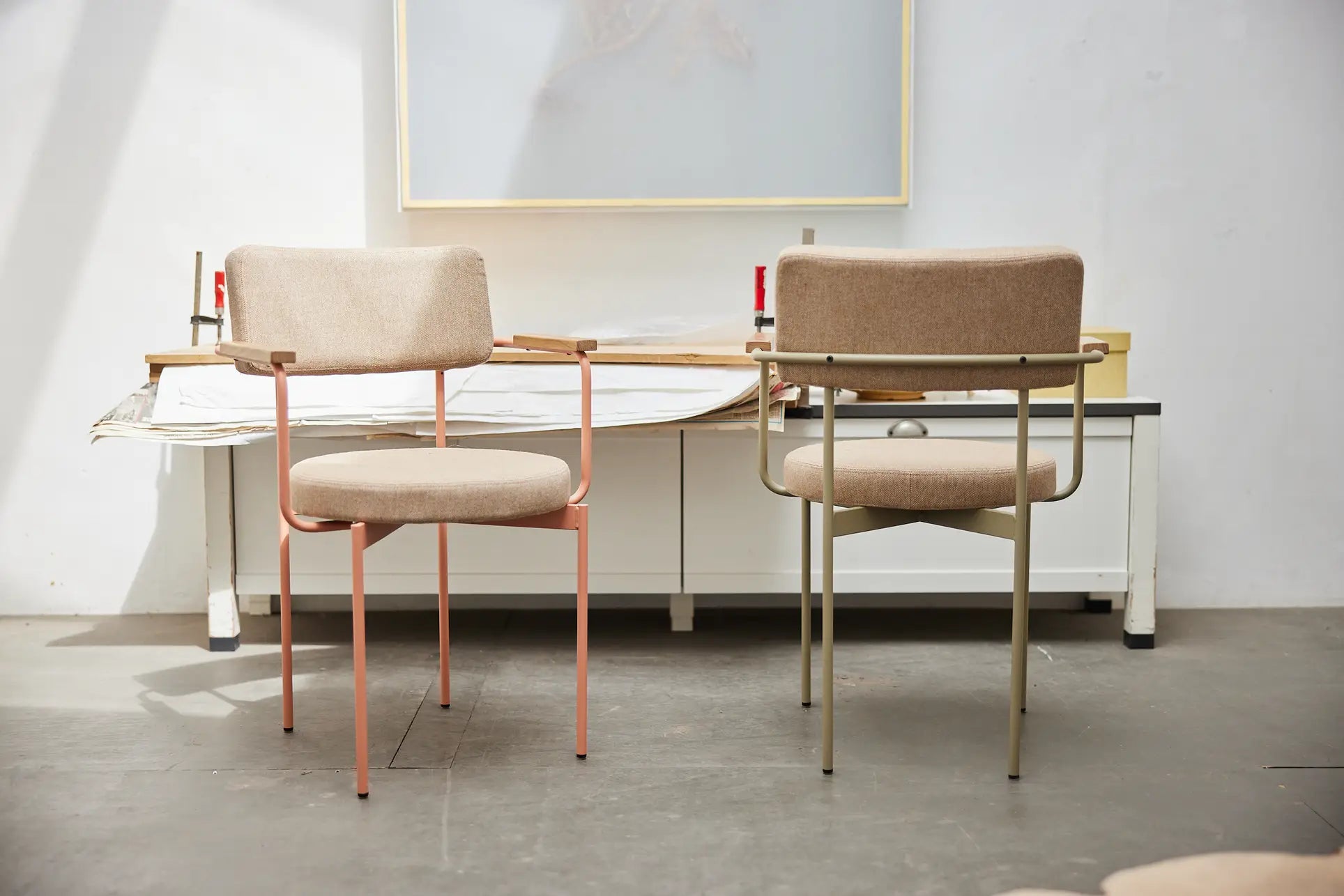 Two modern, stackable HKliving Dining Armchairs in Main Line Flax Nude—one with rose gold and one with brass-colored legs—are paired at a white desk in a bright, minimal room echoing sleek Dutch design.