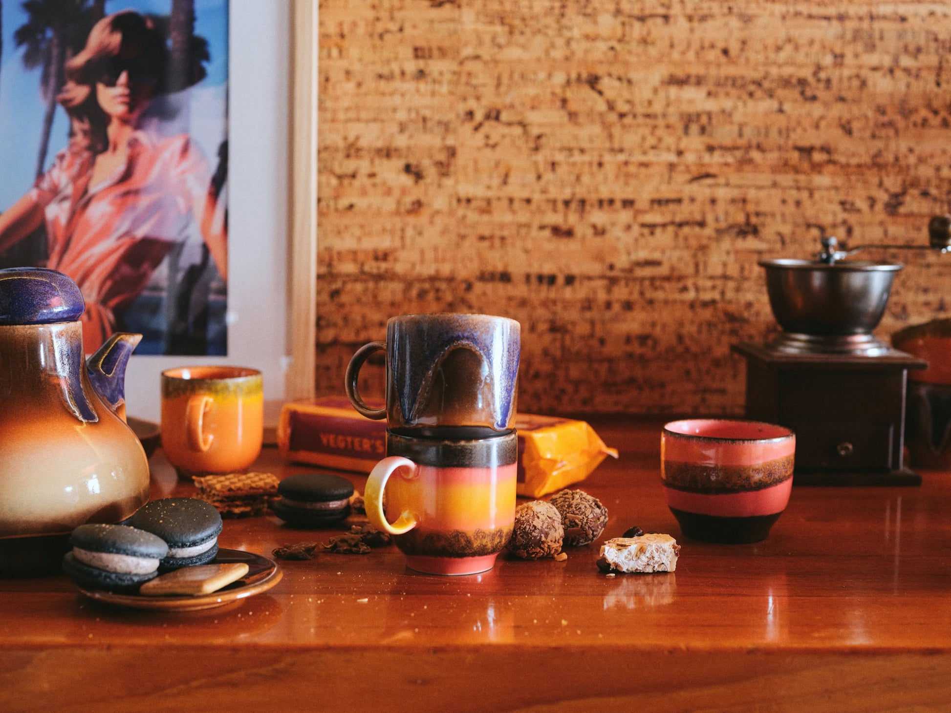 A rustic wooden table features the HKliving 70s Ceramics: Café Mugs (Set of 4), a teapot, assorted cookies, and macarons. A vintage coffee grinder and framed photo add retro charm against a textured wall.
