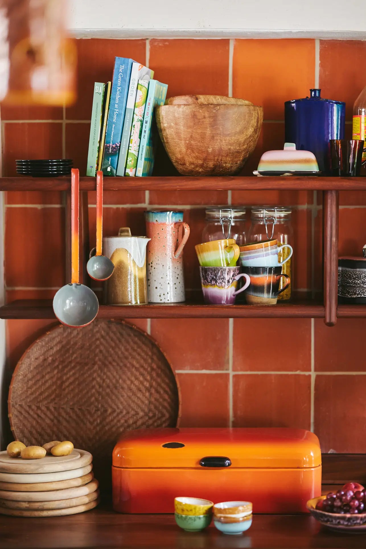 A cozy kitchen shelf with cookbooks, wooden bowls, HKliving 70s Ceramics: Cappuccino Mugs (Set of 4), glass jars, and ladles. Below are plates, a woven mat, potatoes, a red-orange bread box, and small bowls—ideal coffee bar decor.