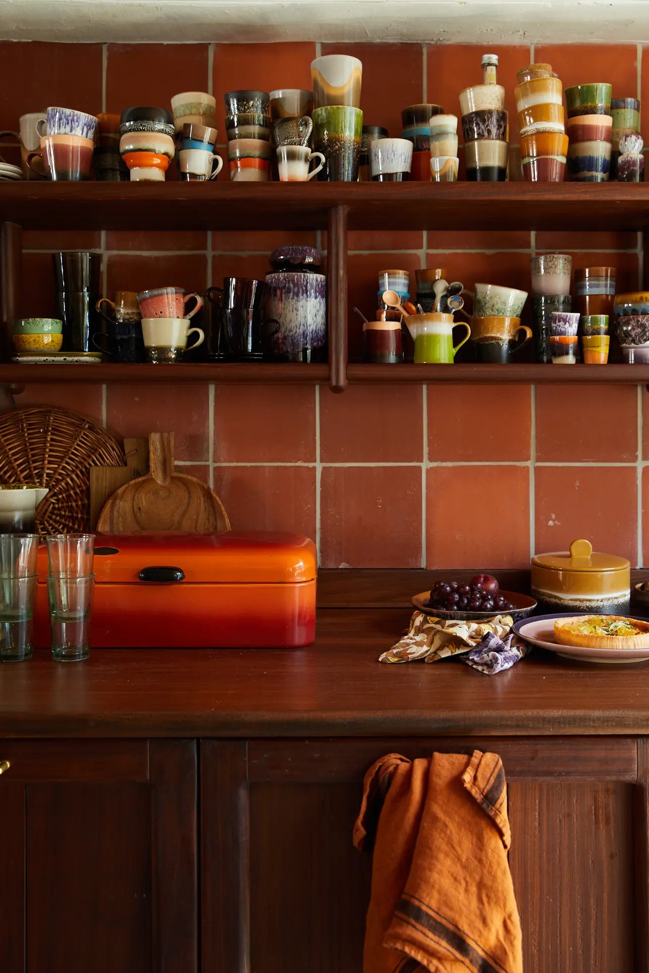 A cozy kitchen with red tiled walls, wooden shelves displaying HKliving 70s Ceramics: Cappuccino Mugs (Set of 4), an orange bread box, glassware, a dish with food, and a rust towel—a perfect coffee bar setup.