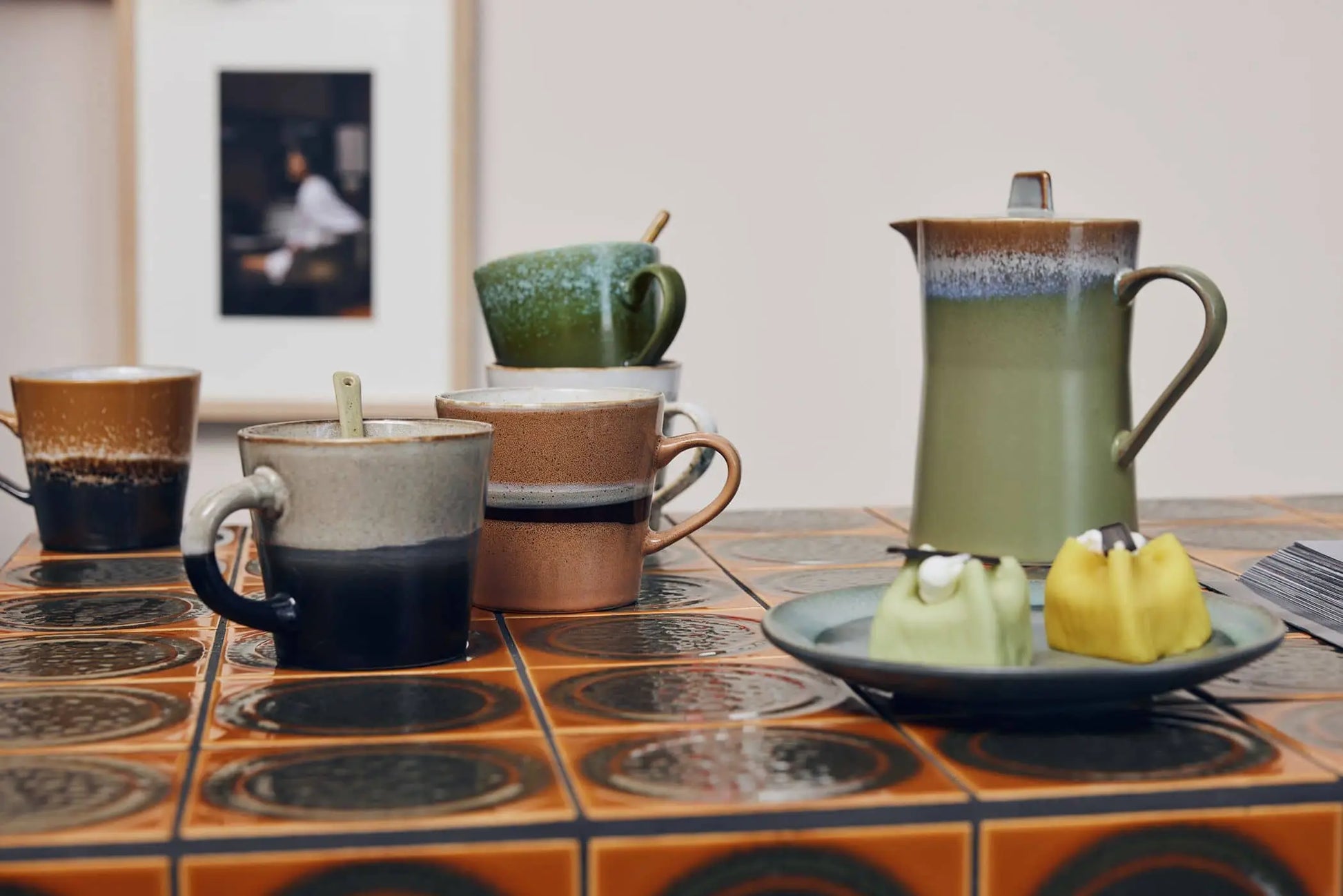A ceramic teapot and a set of HKliving 70s Ceramics: Cappuccino Mugs sit on a patterned table with two green-and-yellow desserts, backed by coffee bar decor and a framed photo on a light-colored wall.
