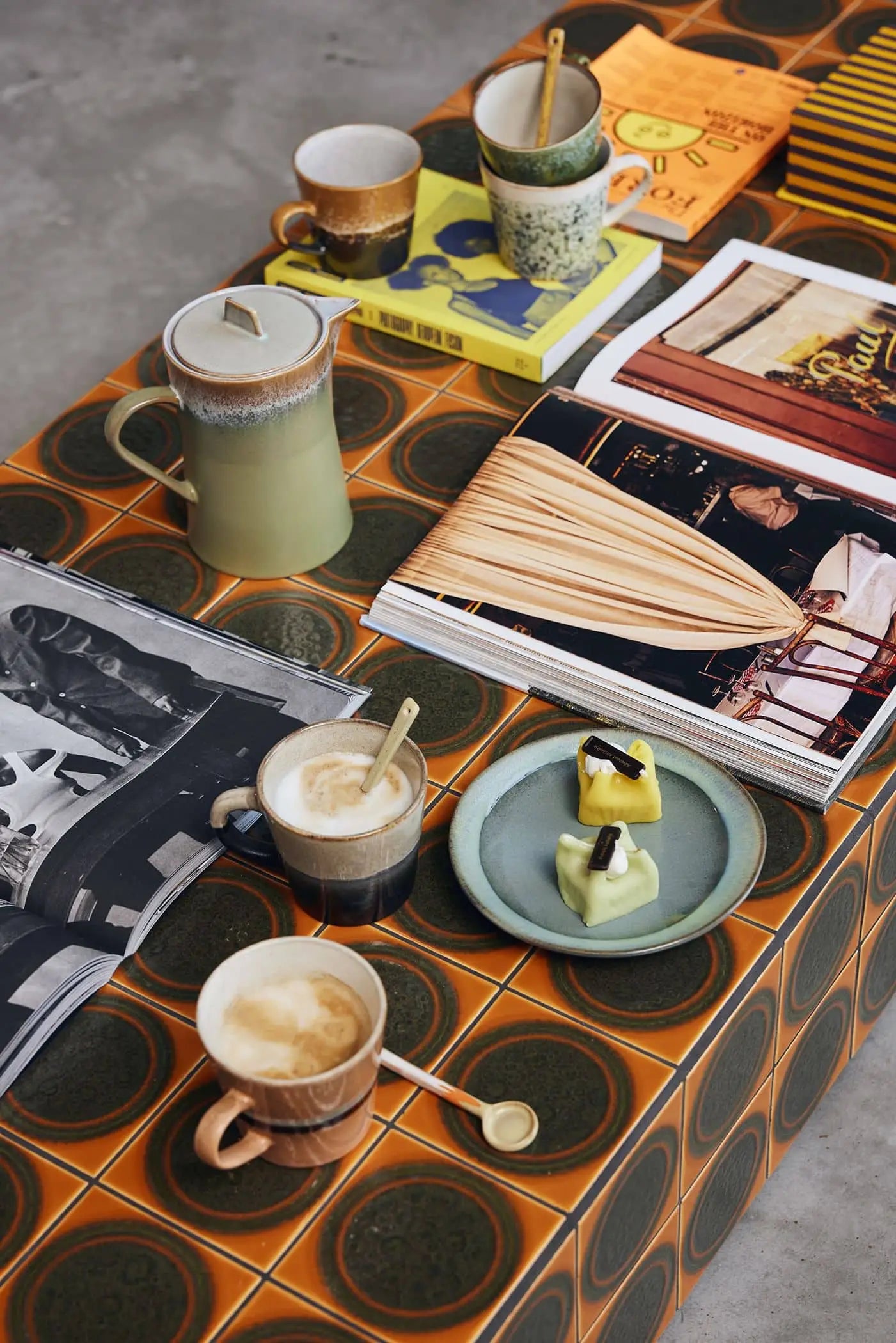 A retro table with a bold orange and green circular pattern displays HKliving 70s Ceramics: Cappuccino Mugs (Set of 4), a ceramic pitcher, pastries on a plate, books, and magazines—ideal for stylish coffee bar decor.