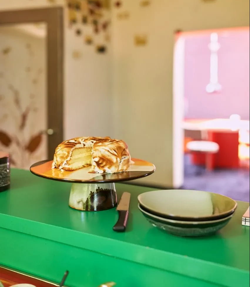 A round cake with a caramelized top sits on a cake stand beside a knife and HKliving 70s Ceramics: Curry Bowls, Set of 2, adding vintage flair to the brightly lit room.