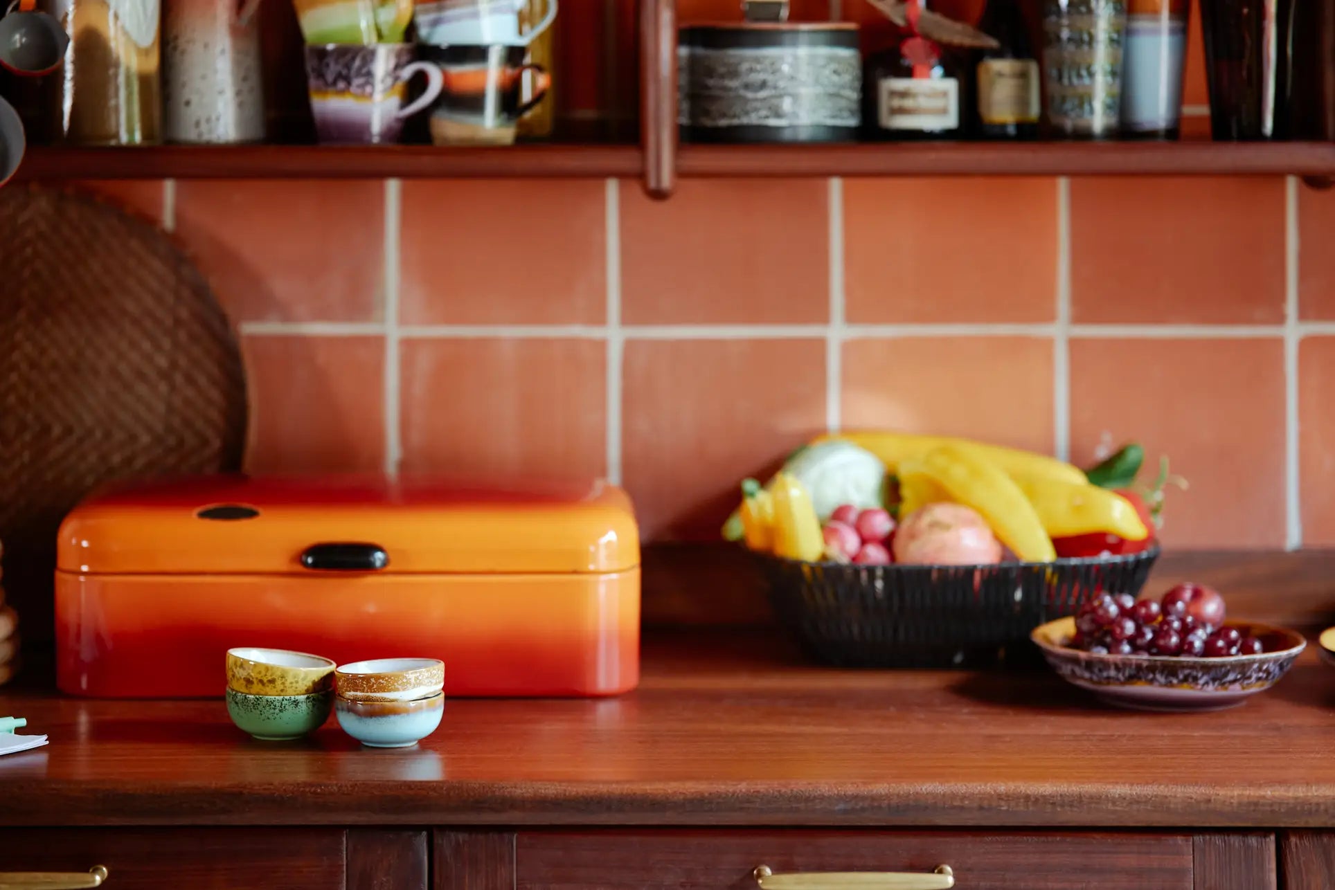 A cozy kitchen countertop with a set of 2 HKliving 70s Ceramics Curry Bowls, an orange bread box, and fresh fruit basket. Above, shelves display mugs and jars, while a warm-toned tile backsplash completes the vintage look.