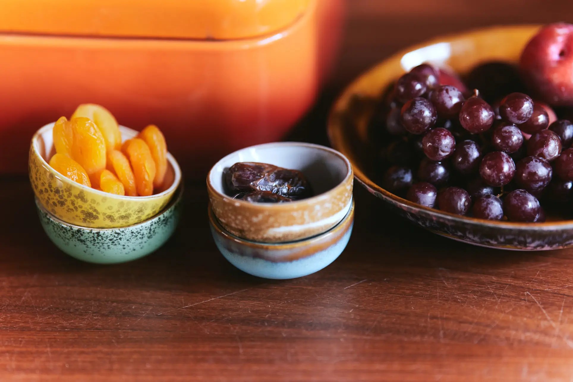 Two HKliving 70s Ceramics Curry Bowls filled with apricots and dates are set next to a larger bowl of grapes and plums on wood, with a pop of 70s style from an orange HKliving dish in the background.