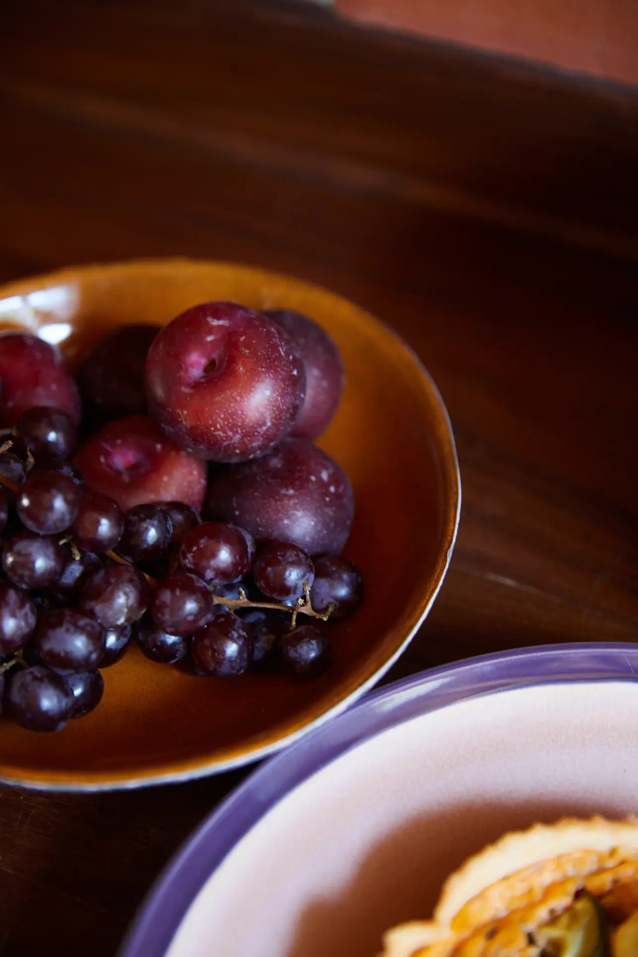 An HKliving 70s Ceramics: Curry Bowl from HKliving, filled with dark red plums and black grapes, sits on wood; in front, a purple-rimmed plate holding a pastry lends vintage flair to the scene.