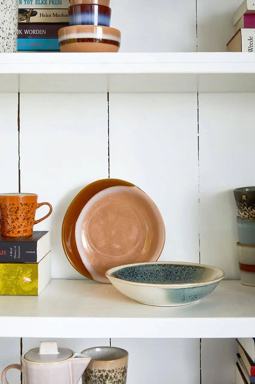 A white shelf displays bowls, plates, mugs, and books. HKliving 70s Ceramics: Curry Bowls (Set of 2) in earthy hues like brown, blue, and beige bring vintage style, while books add extra color and variety to the cozy arrangement.