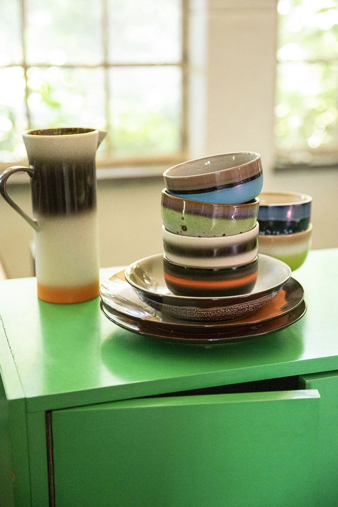 A set of four HKliving 70s Ceramics Dessert Bowls with retro patterns sits on a bright green cabinet in a sunlit room with large windows in the background.
