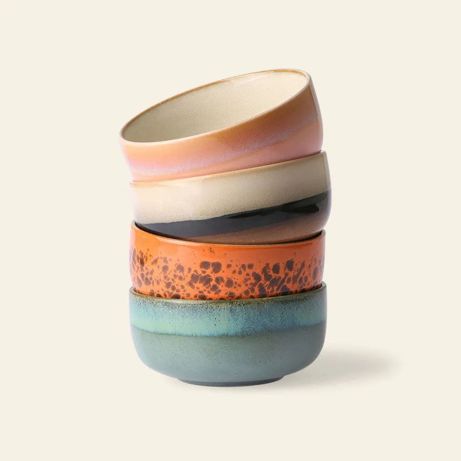 Four HKliving 70s Ceramics Dessert Bowls, each with a unique glaze—pinkish beige, beige with a dark stripe, orange with speckles, and greenish blue—are stacked unevenly against an off-white background.