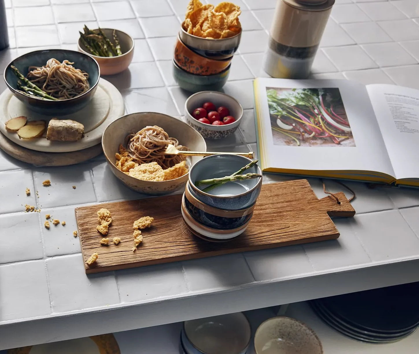 A kitchen table with noodles, cornflakes, cherry tomatoes, ginger, asparagus, an open cookbook on vegetables, and HKliving 70s Ceramics Dessert Bowls (Set of 4) stacked on a wooden board atop the white tiled surface.