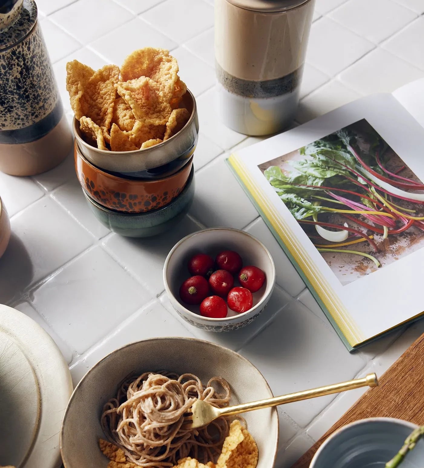 A set of HKliving 70s Ceramics Dessert Bowls filled with cherry tomatoes and snacks, alongside retro ceramic cups, noodles with a fork, and an open cookbook on leafy vegetables arranged on a white tiled surface.