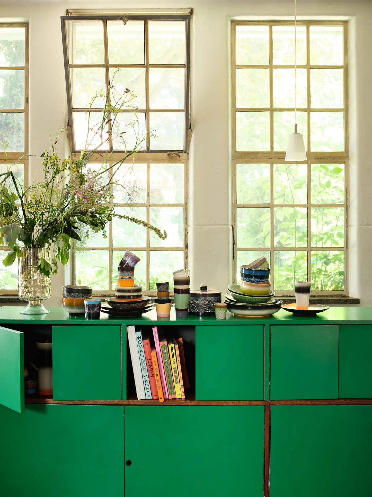 A bright kitchen with green cabinets, stacked dishes and bowls, colorful books on an open shelf, and sunlight streaming in—perfect for a morning espresso in the HKliving 70s Ceramics: Latte Mugs, Set of 2 by HKliving.