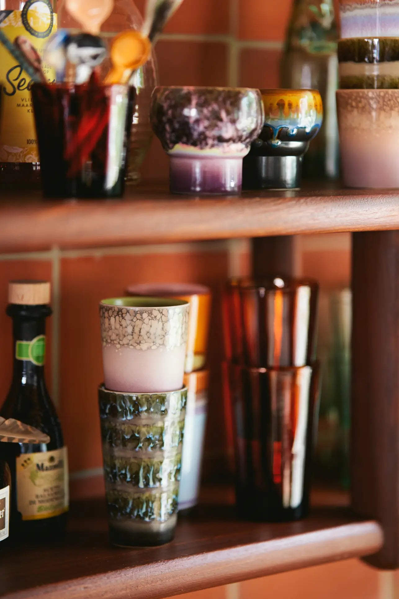 HKliving 70s Ceramics: Latte Mugs, Set of 2 by HKliving are displayed among glass cups and bottles on wooden shelves against a tiled wall, their unique textures and patterns adding to the eclectic arrangement.