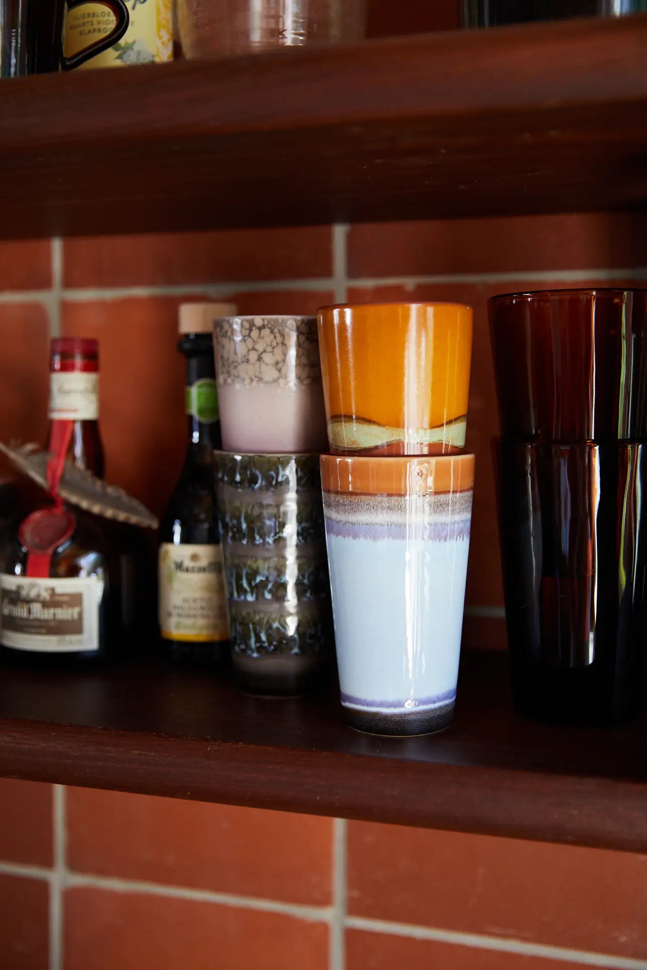 A wooden shelf displays HKliving 70s Ceramics Latte Mugs (Set of 2) alongside clear glasses and colorful cups, all arranged against a red tiled wall.