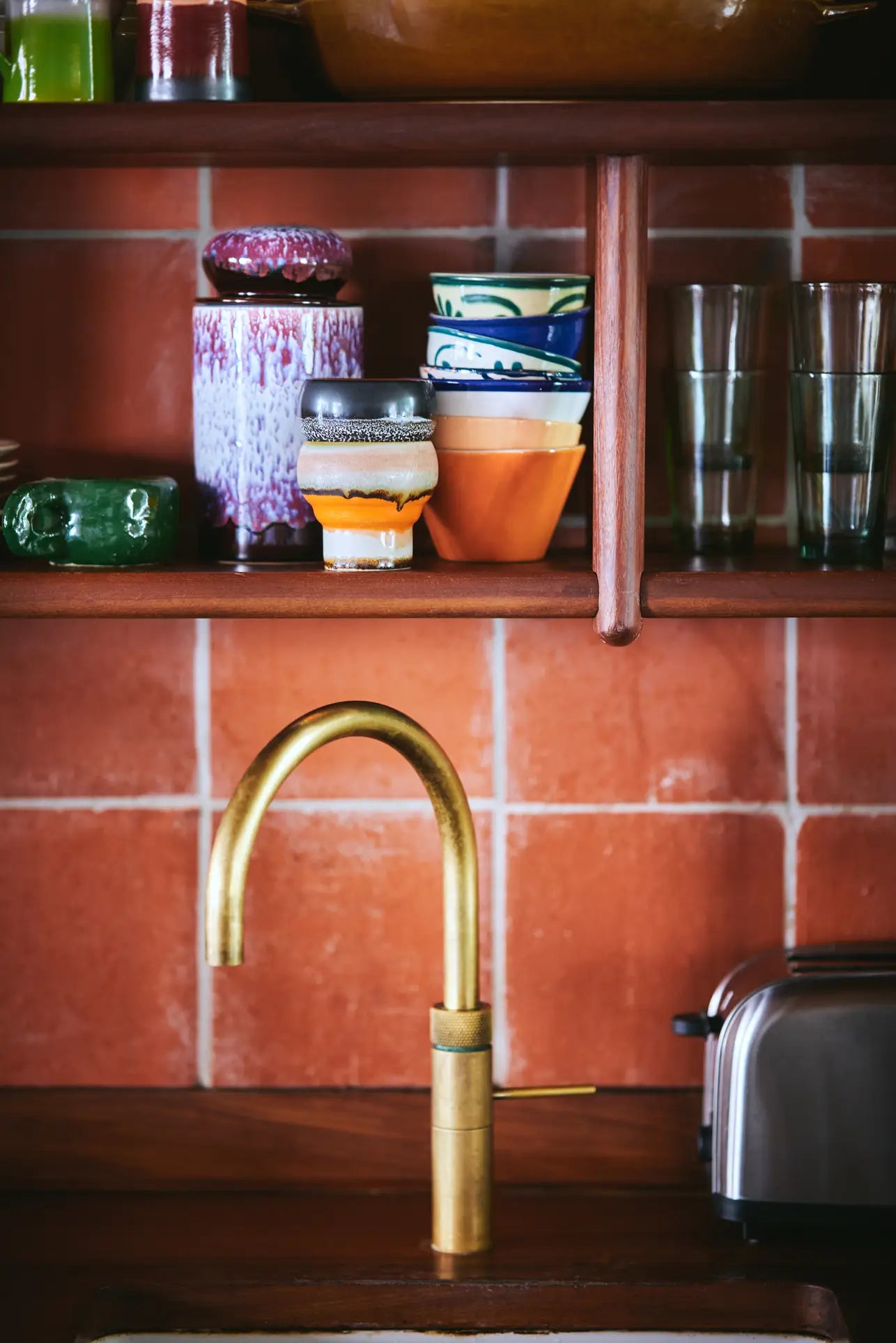 A brass kitchen faucet sits below wooden shelves displaying HKLIVING 70s Ceramics Lungo Mugs—perfect for coffee lovers. Nearby, a metallic toaster rests against the terracotta tile backsplash.