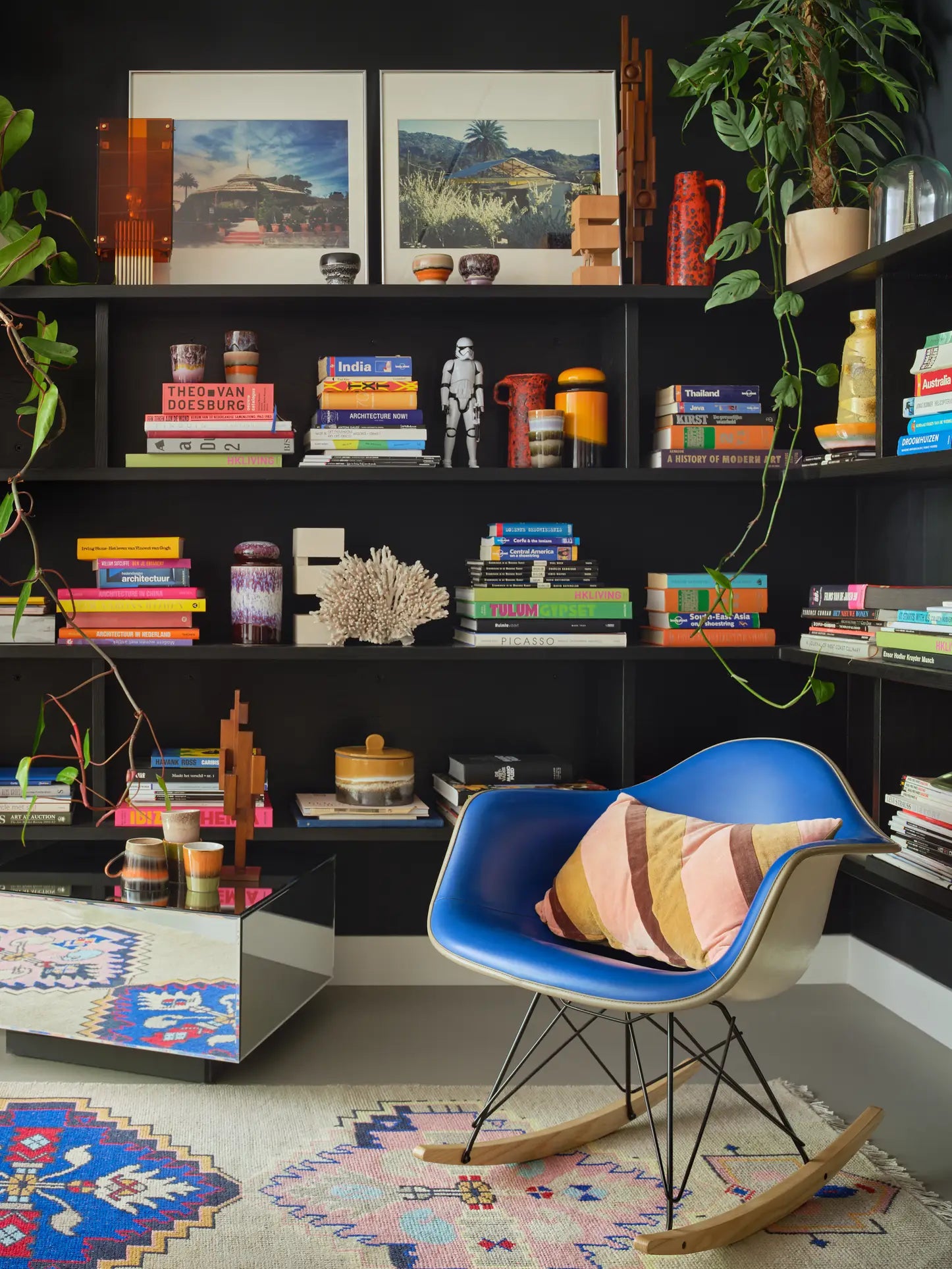 A cozy reading nook with a blue rocking chair, striped pillow, colorful rugs, bookshelves filled with books, plants, vases, HKLIVING 70s Ceramics: Lungo Mugs (Set of 2), art prints, and décor against a dark wall.