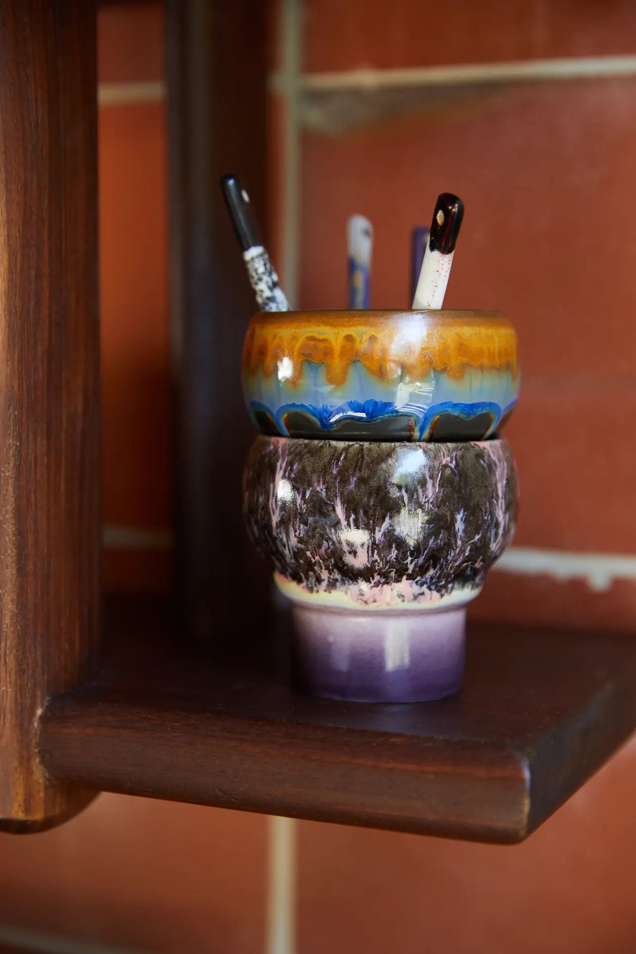 Two HKLIVING 70s Ceramics Lungo Mugs (Set of 2) sit stacked on a wooden shelf, filled with paintbrushes against a red tile wall—a vibrant scene for coffee and art lovers.