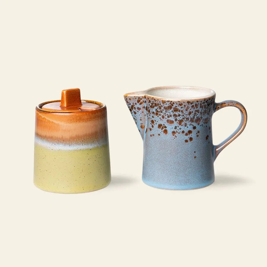 The HKliving 70s Ceramics: Milk Jug & Sugar Pot set features earthy brown, green, and blue tones with a speckled glazed finish by HKliving, displayed against a light beige background.