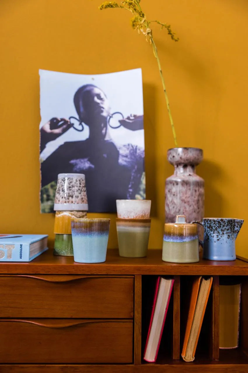 HKliving 70s Ceramics milk jug and sugar pot sit on a wooden cabinet. Behind them, a black-and-white photo of a person with large hoop earrings is pinned to a mustard yellow wall, with books placed on and inside the cabinet.