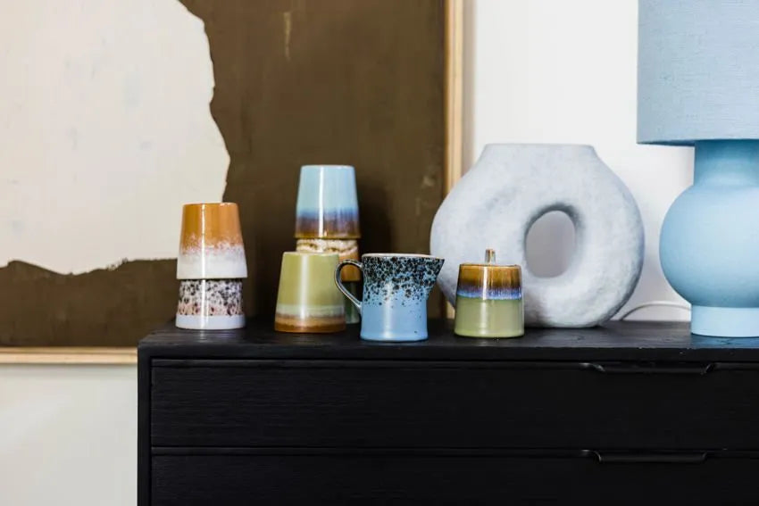 A modern black dresser displays HKliving 70s Ceramics: Milk Jug & Sugar Pot, a blue speckled mug, and a round white textured vase, while a blue lamp adds charm against abstract wall art.