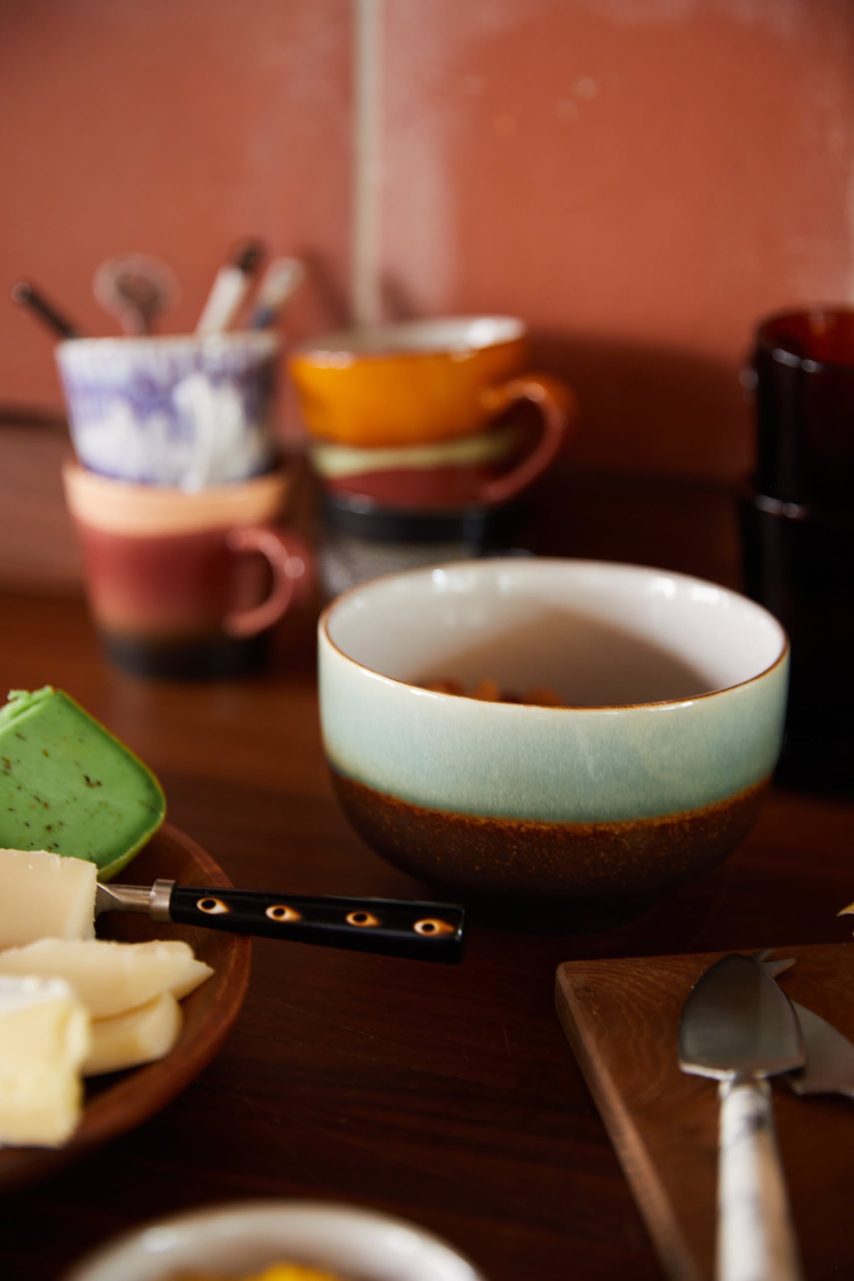 A close-up of HKliving 70s Ceramics Noodle Bowls, Set of 4, on a wooden table with slices of cheese and colorful mugs in the background—perfect for serving your favorite dishes.
