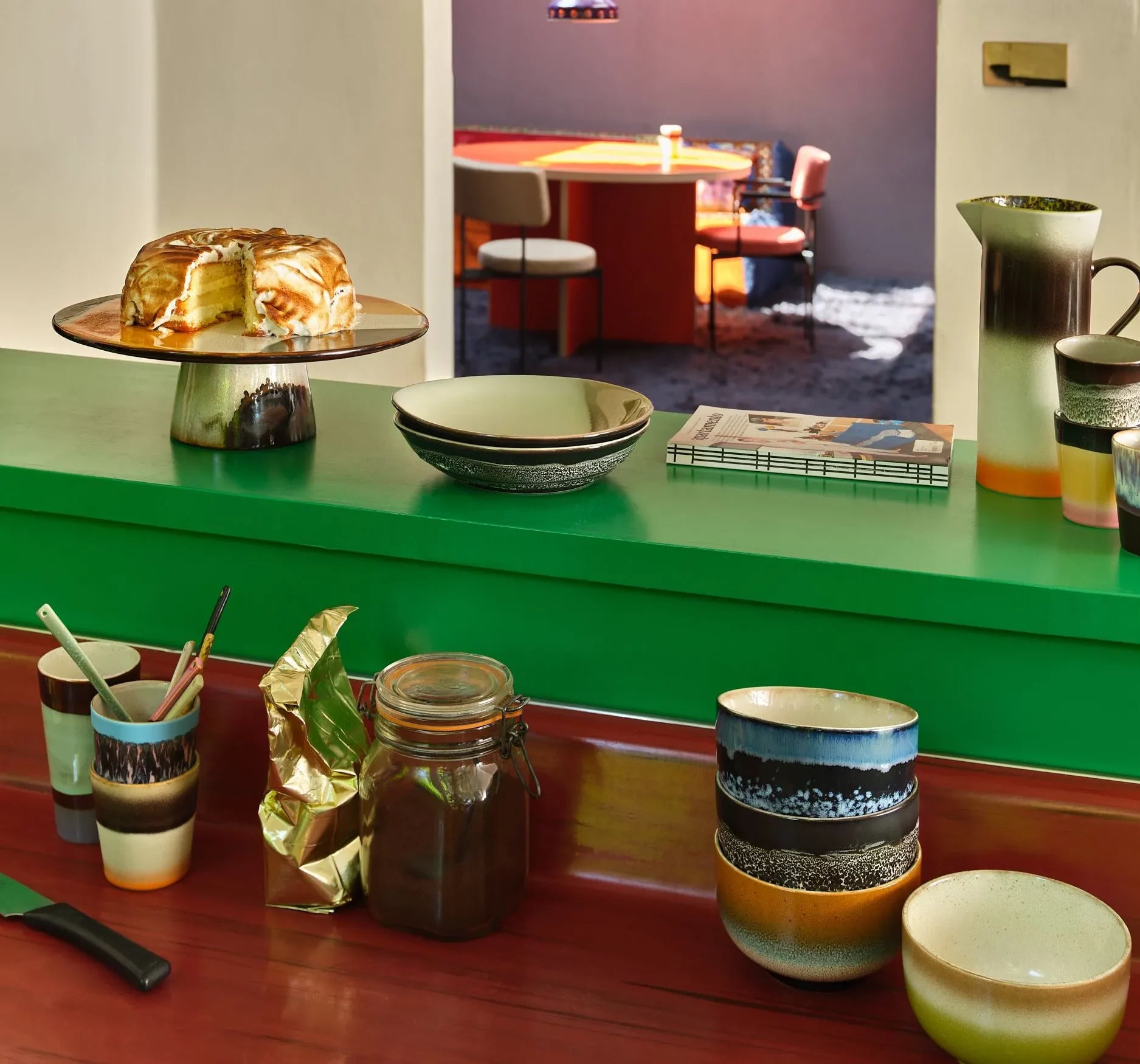 A kitchen counter features HKliving 70s Ceramics Noodle Bowls (set of 4), cups, utensils, a jar, and a cake stand. In the background, sunlight fills a dining area with a red table and chairs—ideal for sharing your favorite meals.