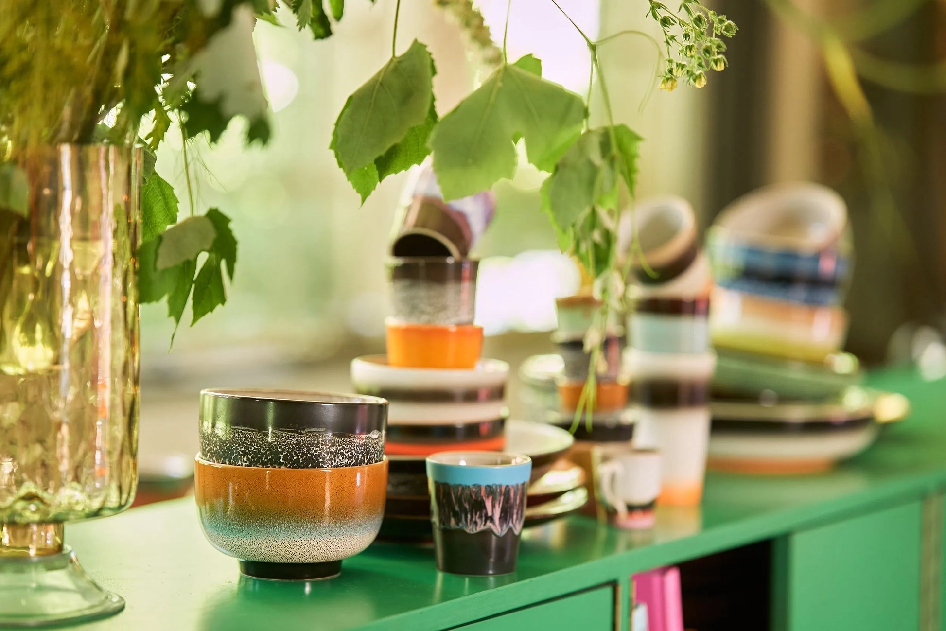 HKliving 70s Ceramics Noodle Bowls, Set of 4, are stacked on a green cabinet beneath leafy plants. Sunlight streams through the window, creating an inviting spot to enjoy your favorite meals.