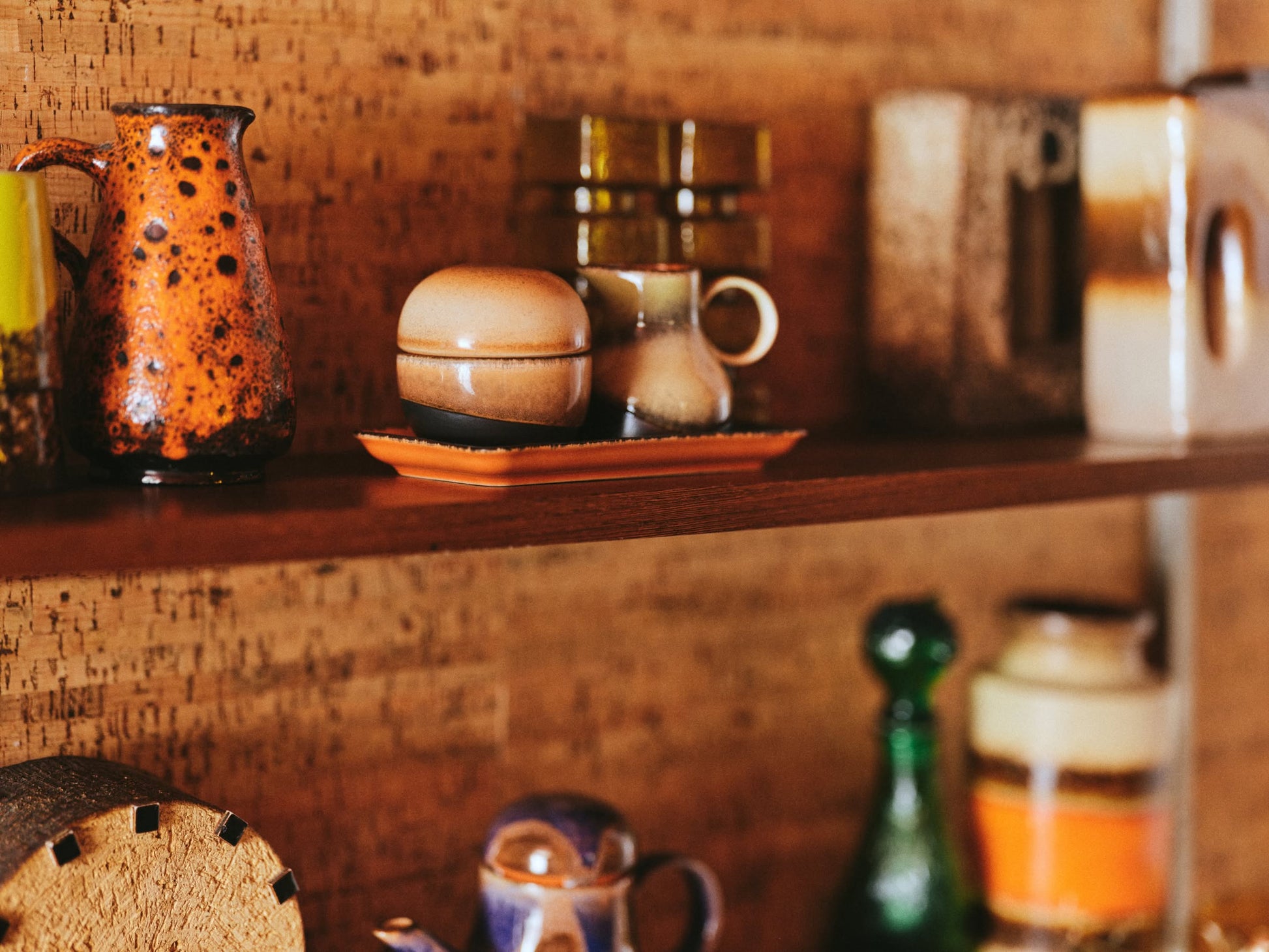 A wooden shelf showcases vintage ceramics, featuring the HKliving 70s Ceramics: Sugar Pot by HKliving for condiments, alongside a speckled orange pitcher, cups, glass bottles, and assorted pottery set against a textured background.