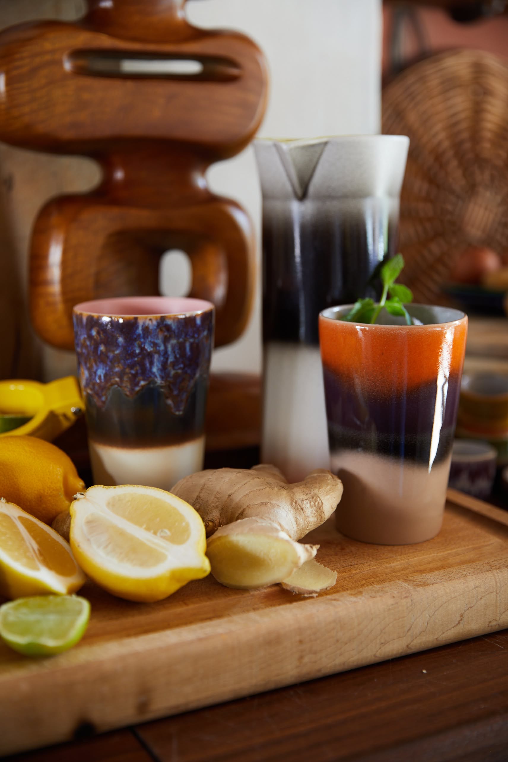The HKliving 70s Ceramics Tea Mugs, Set of 2, are displayed on a wooden board with lemon, lime, ginger, and mint; a matching pitcher and woven basket in the background add retro flair to this vibrant tea setting.