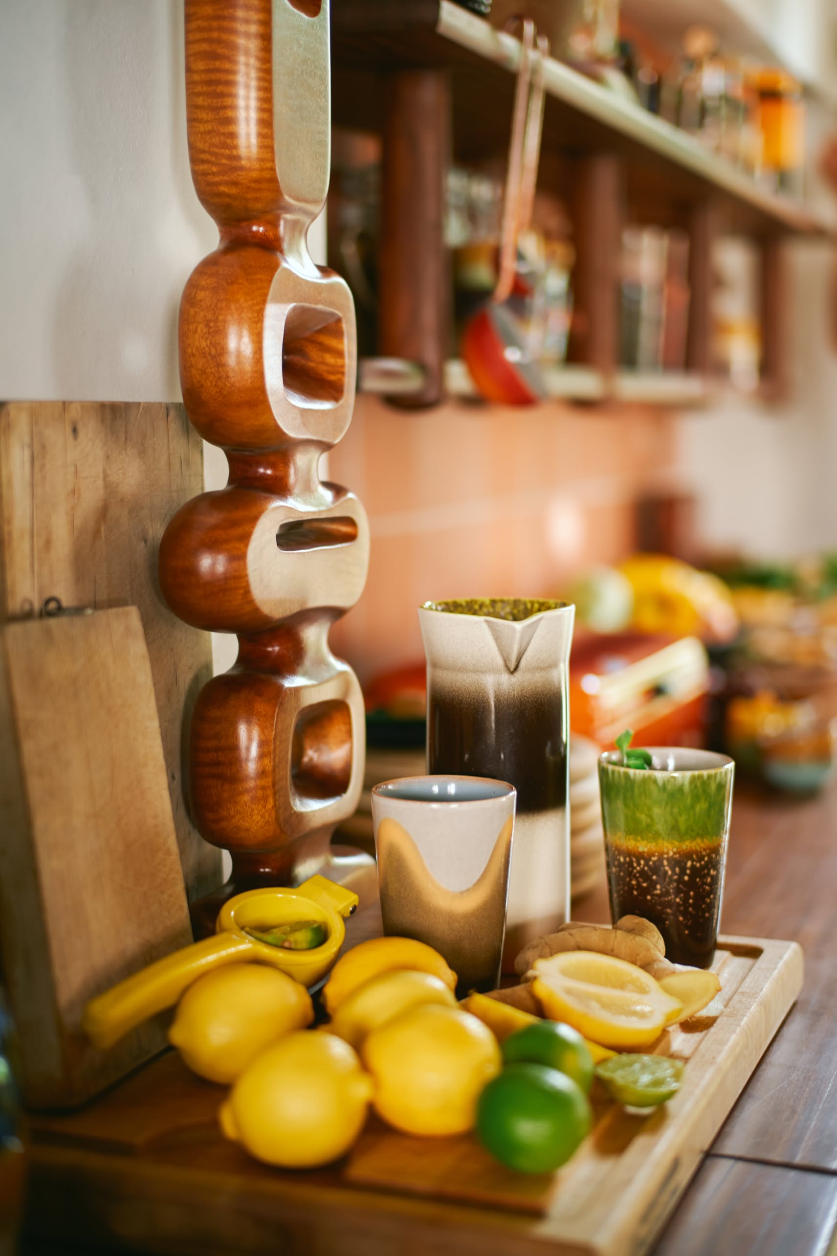 A wooden cutting board holds lemons, a lime, a banana, HKliving 70s Ceramics tea mugs (set of 2), and a glass with a drink. A modern wooden sculpture and a kitchen shelf with retro ceramics and jars create a unique background.