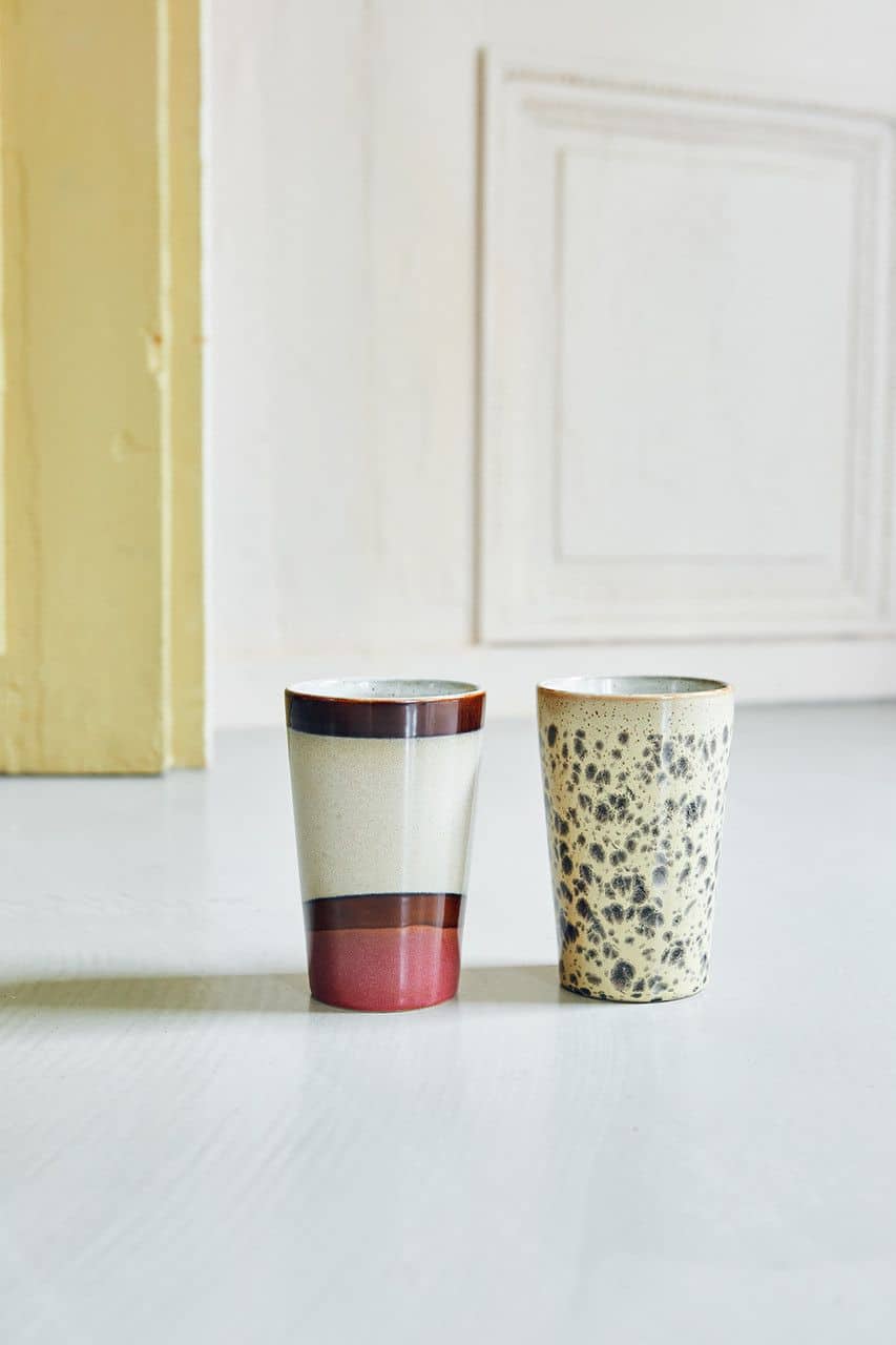 Two HKliving 70s Ceramics Tea Mugs sit on a light surface—one features cream, brown, and rust-red bands for a vintage look, while the other is beige with a mottled brown speckle. Pale walls enhance the retro 70s Ceramics vibe.