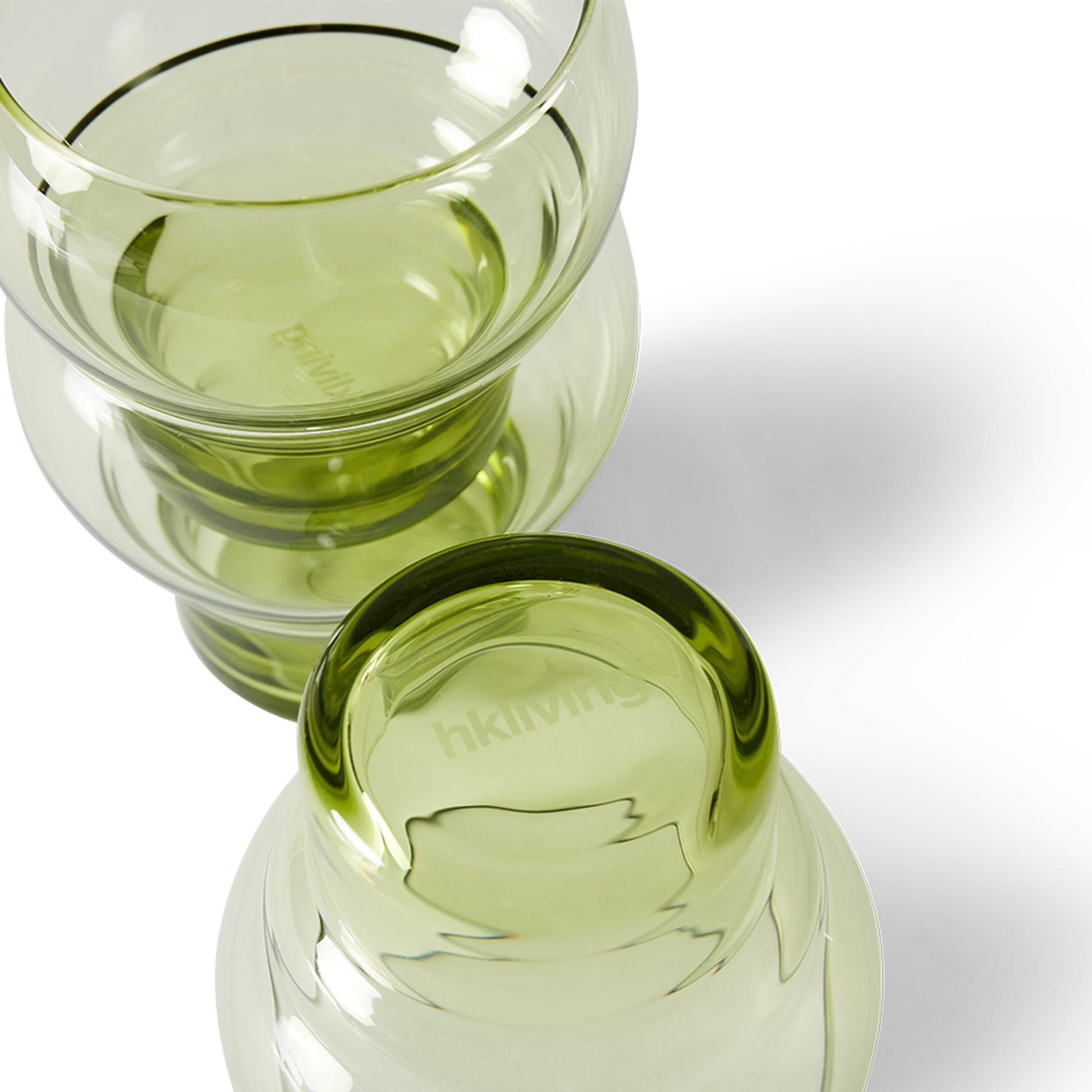 A set of four translucent, light green HKliving 70s Glassware Bulb Glasses is stacked on a white surface with soft shadows and the HKliving logo on the base. Also available in amber.