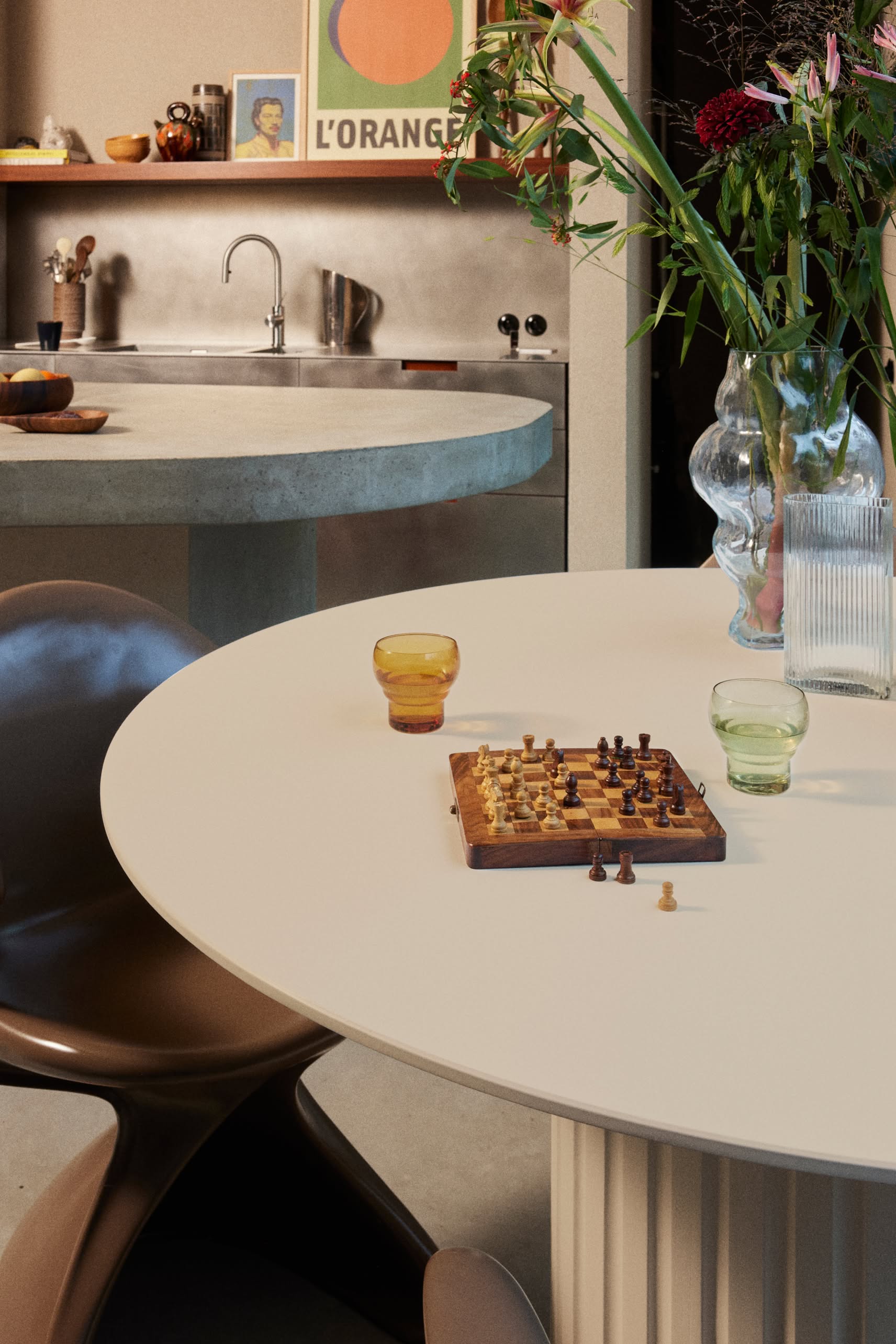 A modern kitchen features a round white table with a chessboard, HKliving 70s Glassware: Bulb Glasses (Set of 4), and flower vases. In the background, a sink and a framed “L’ORANGE” poster lend retro sophistication to the room.