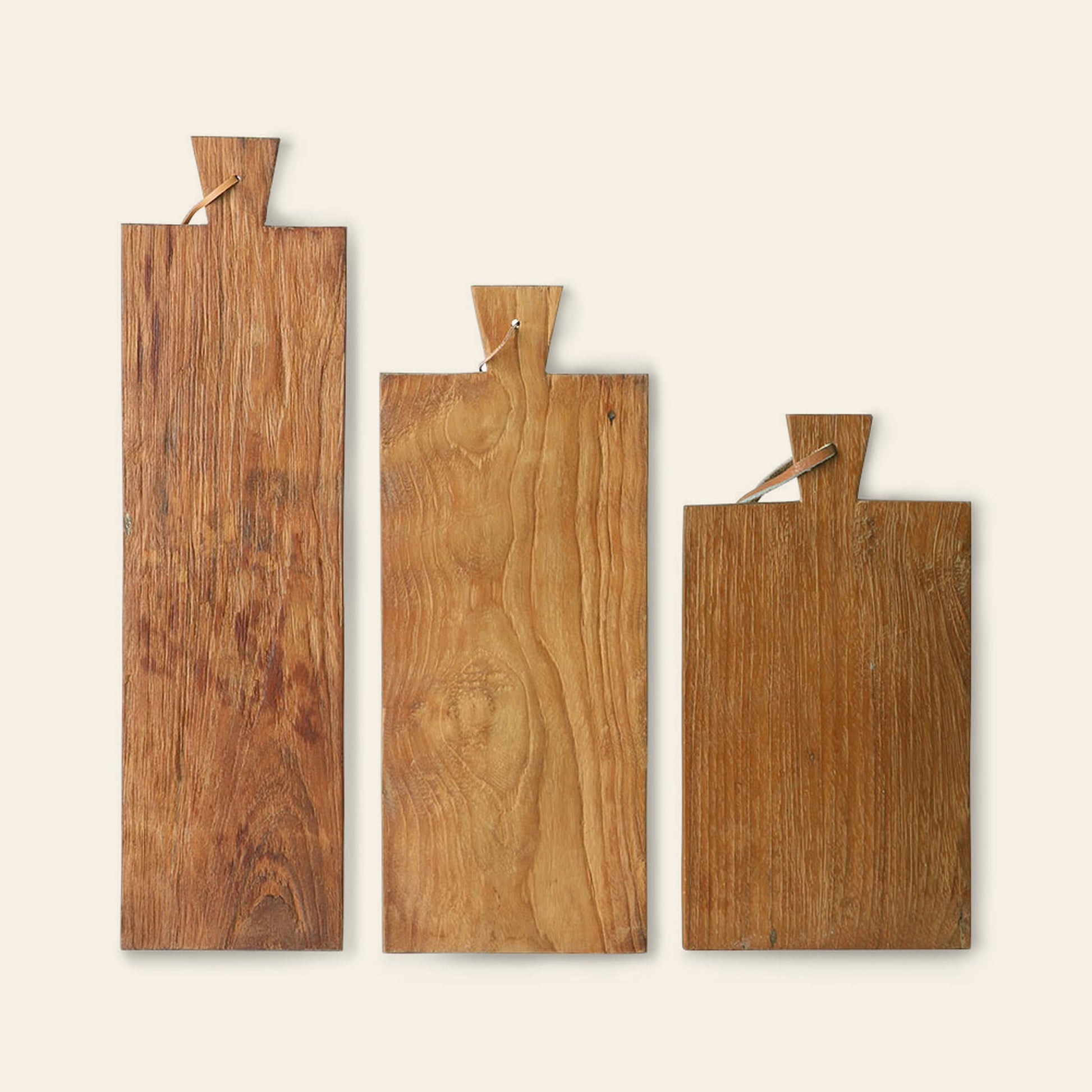 The HKliving Bread Board Teak, Set of 3 by HKliving features three teak bread boards in assorted sizes with handle cutouts and string loops for hanging, displayed upright to showcase rustic charm against a light cream background.