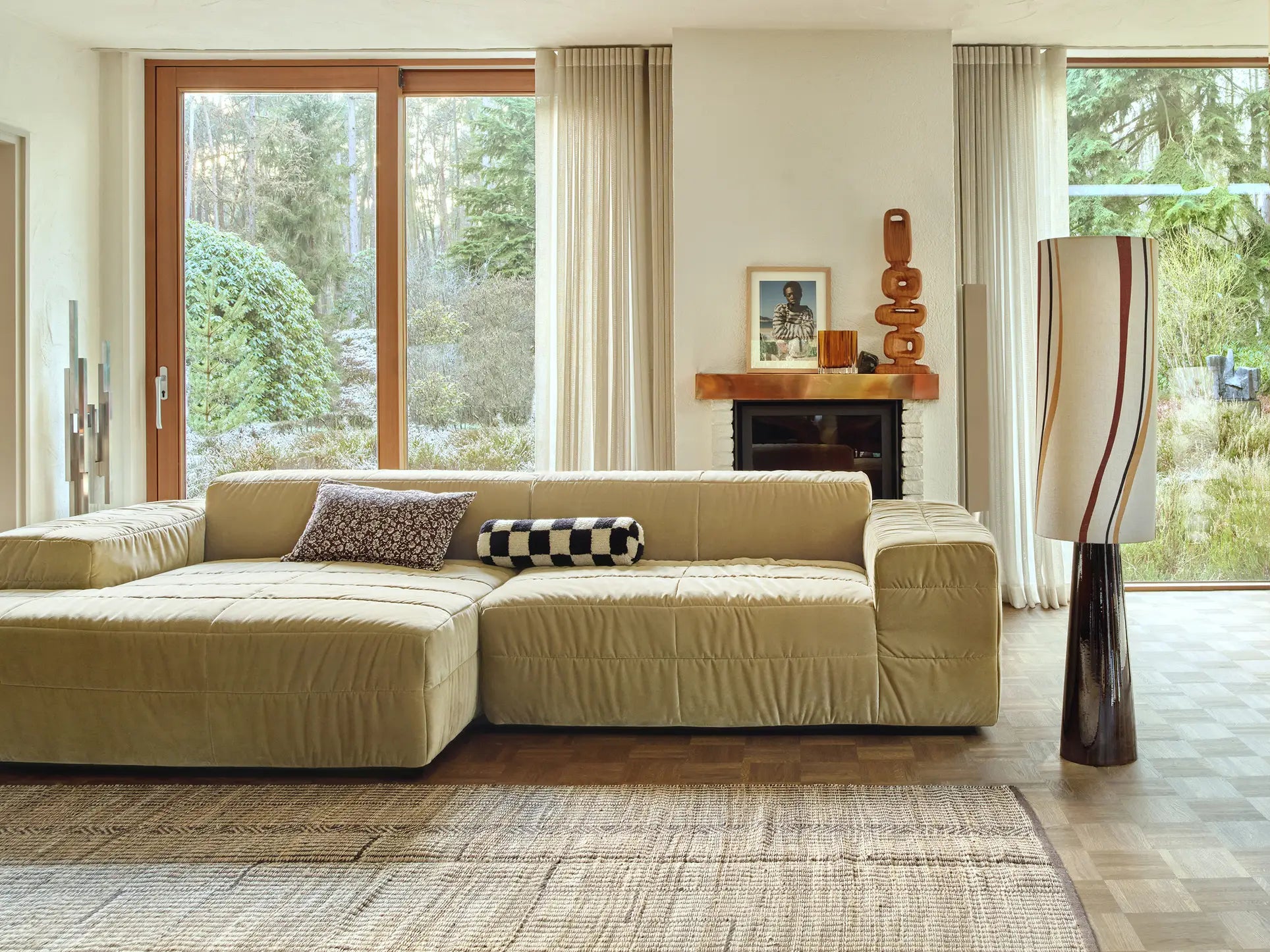 A modern living room features the HKliving Brut Sofa, Left by HKliving—a beige sectional with patterned pillows, a textured rug, tall windows with sheer curtains, a stylish fireplace, and views of a lush wooded garden.