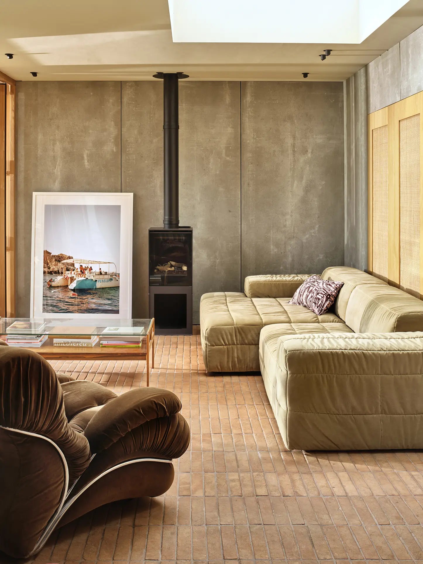 A modern living room with spacious seating, featuring the HKliving Brut Sofa, Right Divan in tan, a brown vintage lounge chair, wood-burning stove, glass coffee table, and a framed boat photo against a concrete wall on a brick floor.