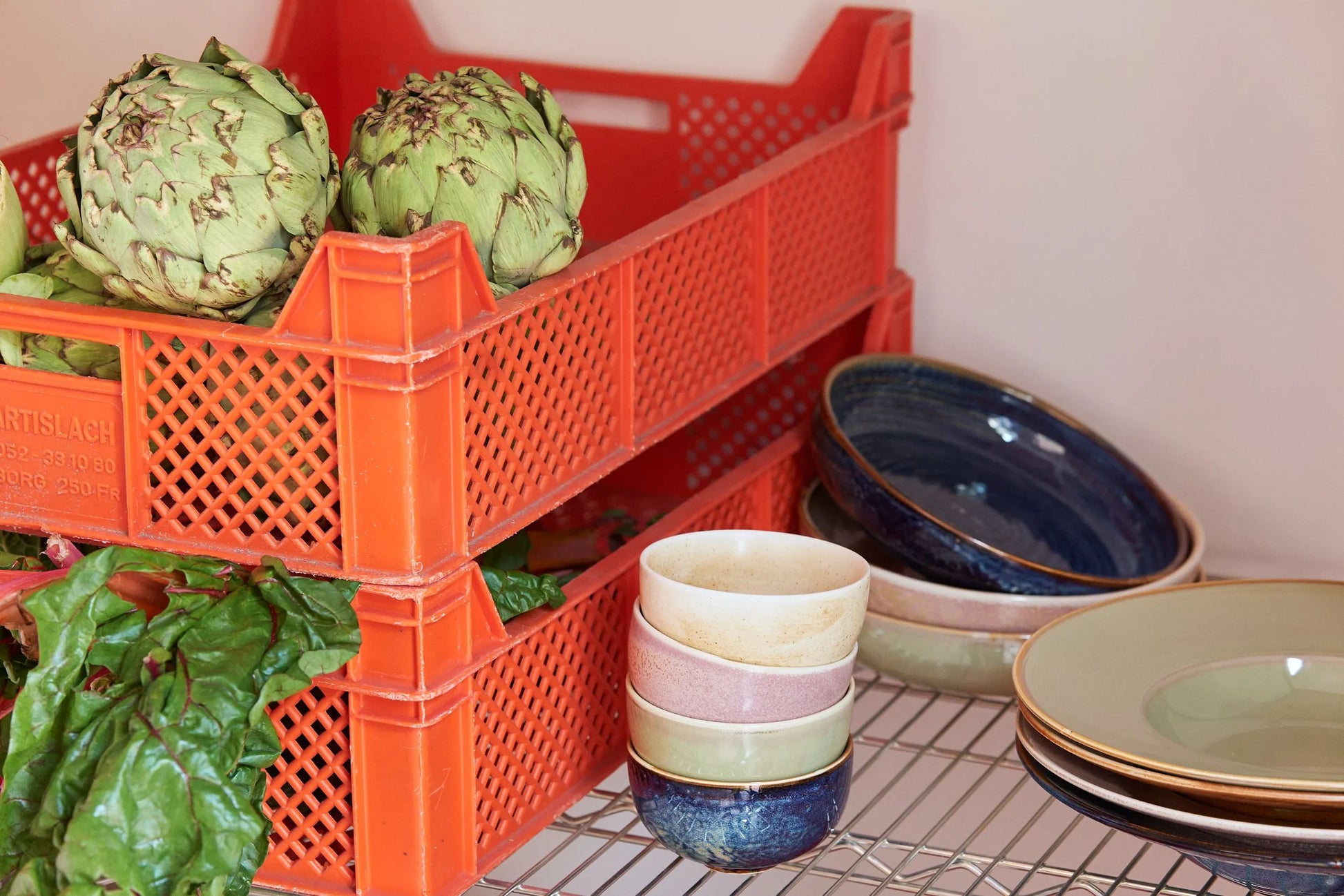 Two stacked orange crates filled with artichokes and greens sit by a wire shelf displaying HKliving Chef Ceramics bowls—vibrant, reactive-glazed pieces by HKliving—bringing color to the softly lit scene and highlighting tableware essentials.