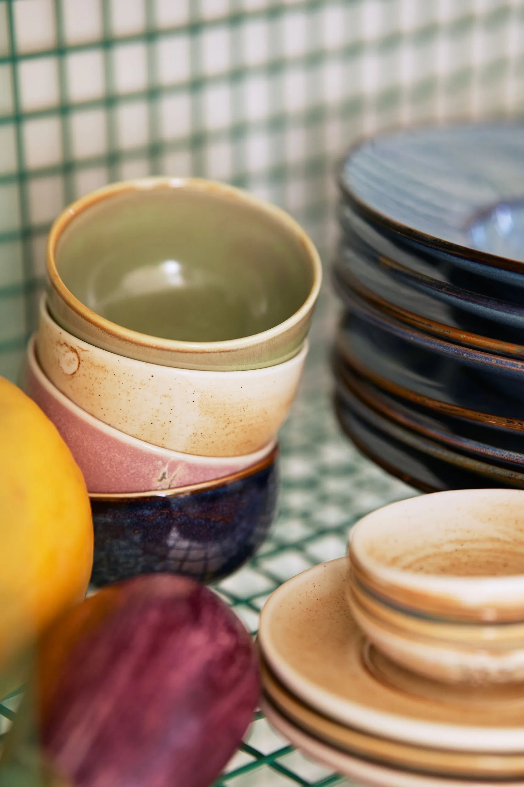 A stack of three HKliving Chef Ceramics Bowls sits beside blue-rimmed plates and saucers, accented by a lemon and purple fruit—stylish HKliving tableware on a green grid-patterned surface.