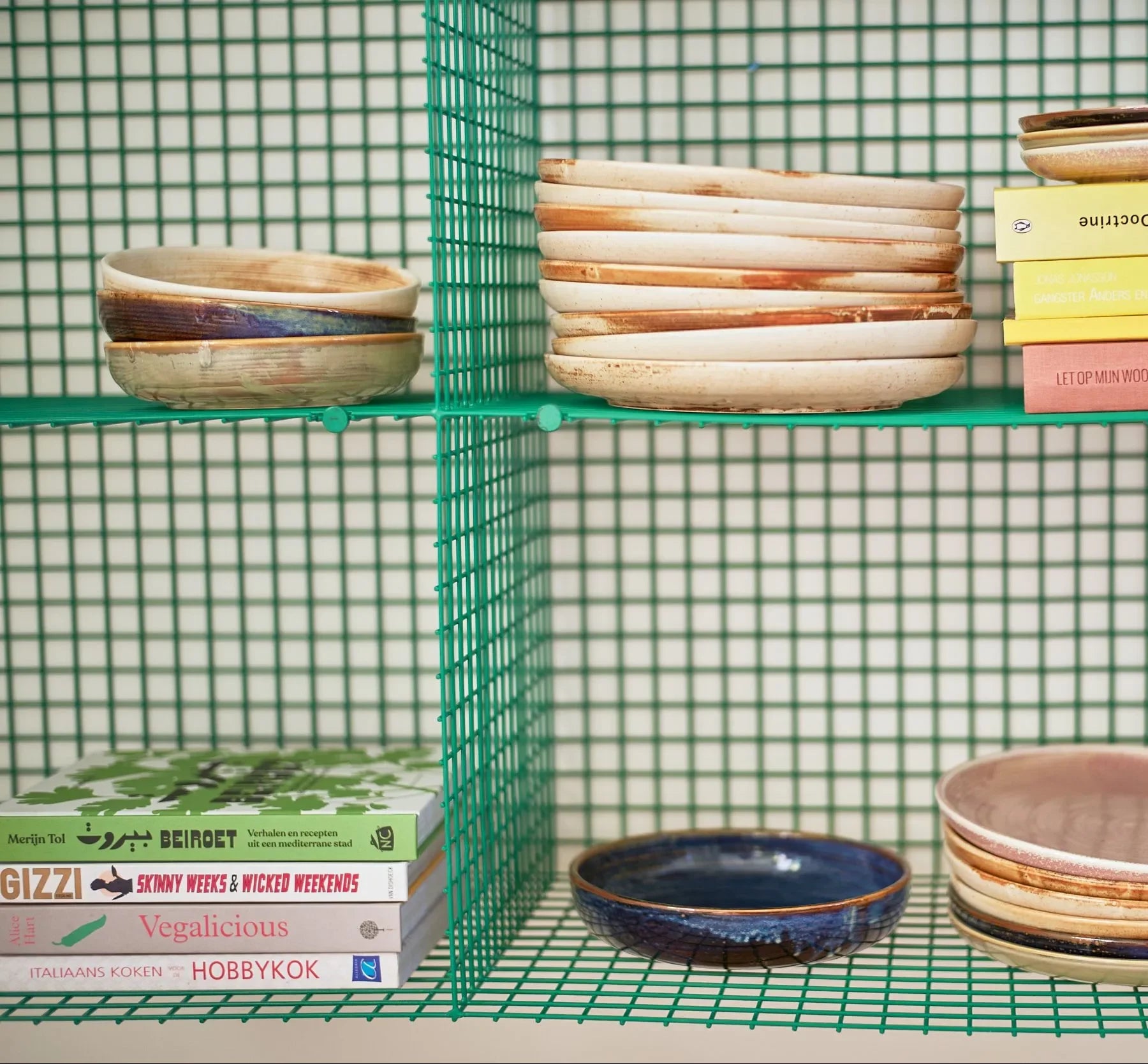 HKliving Chef Ceramics: Deep Plate M by HKliving, in assorted colors, is stacked on green wire shelves. Nearby, several cookbooks are displayed against a green grid-patterned backdrop.