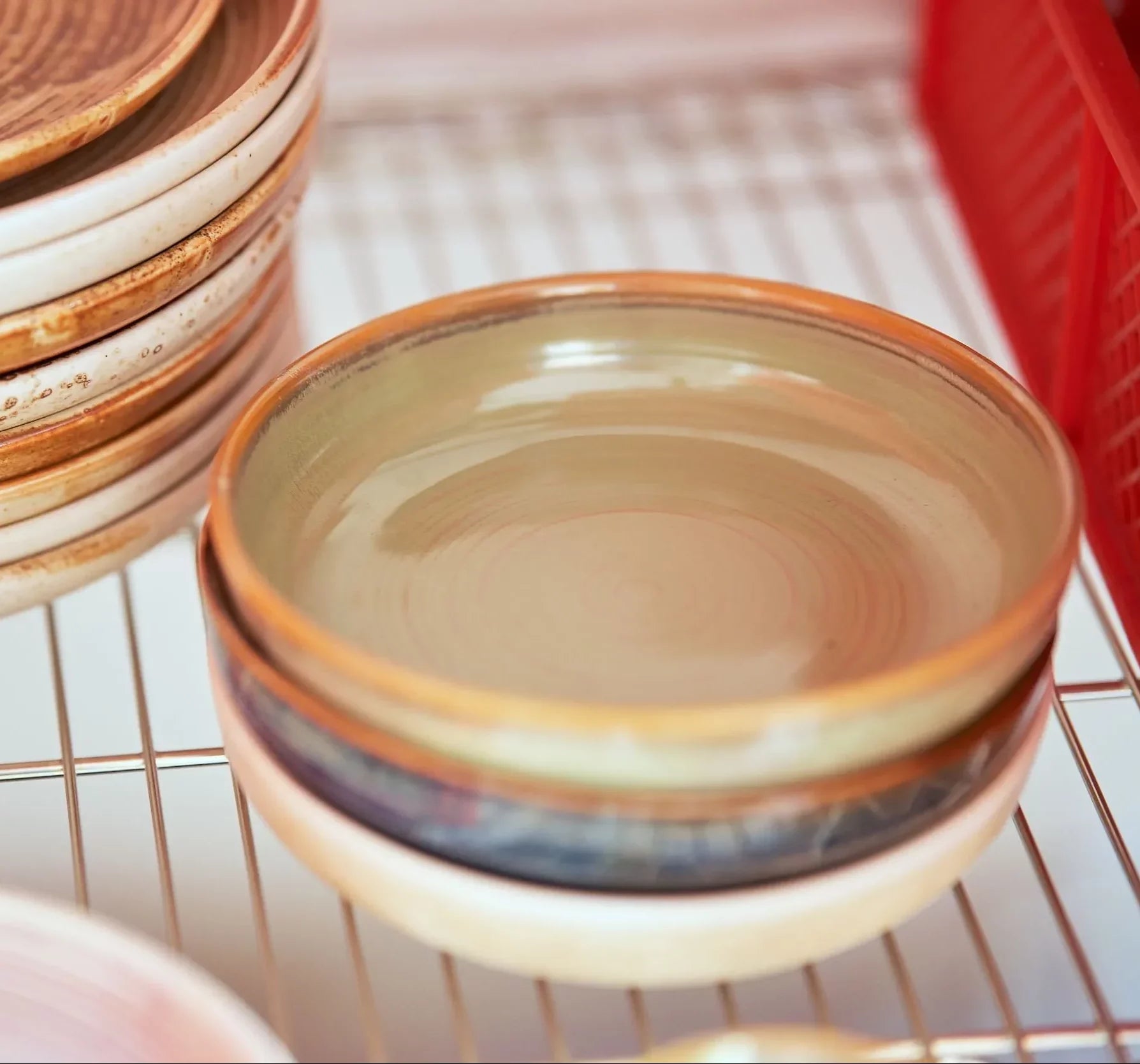 Four HKliving Chef Ceramics: Deep Plate M by HKliving are stacked on a dish rack, with more plates to the left and a red plastic rack to the right. Their earthy tones and reactive glaze create an artisanal look.